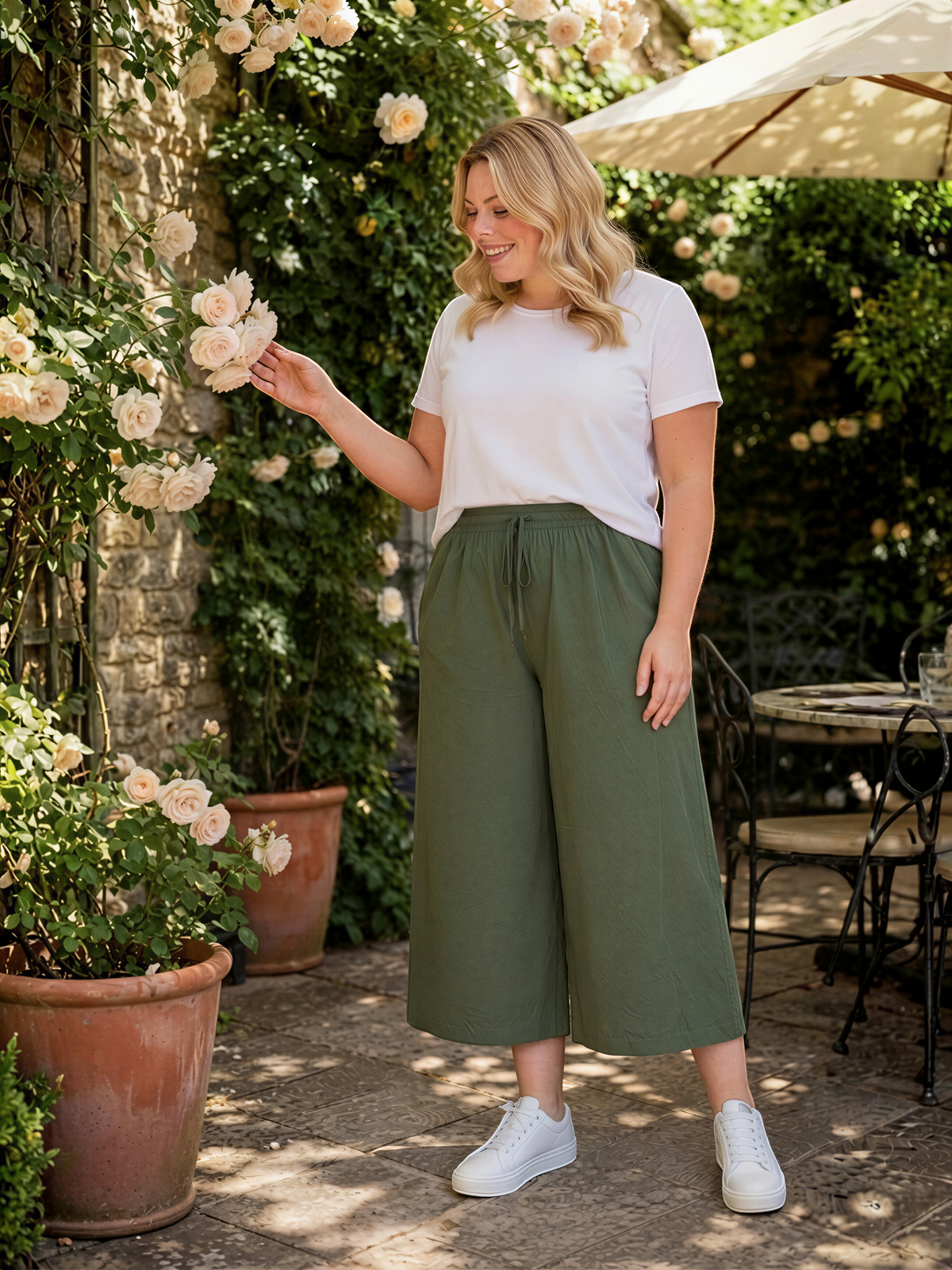 A woman in a white t-shirt and olive green wide-leg pants stands in a garden, holding a flower and smiling.