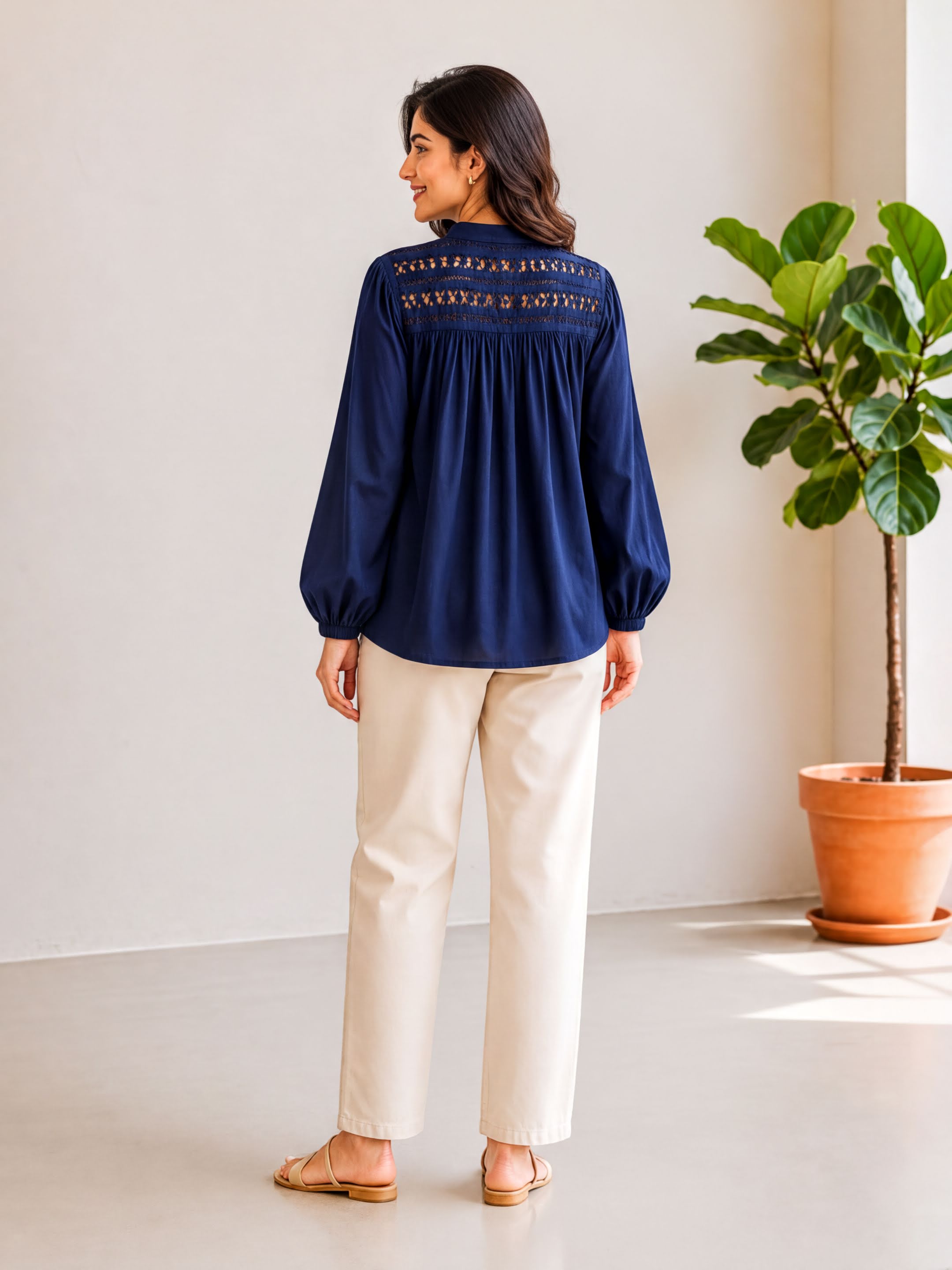 A woman wearing a navy blue blouse with embroidered designs on the back stands in a room with a potted plant.