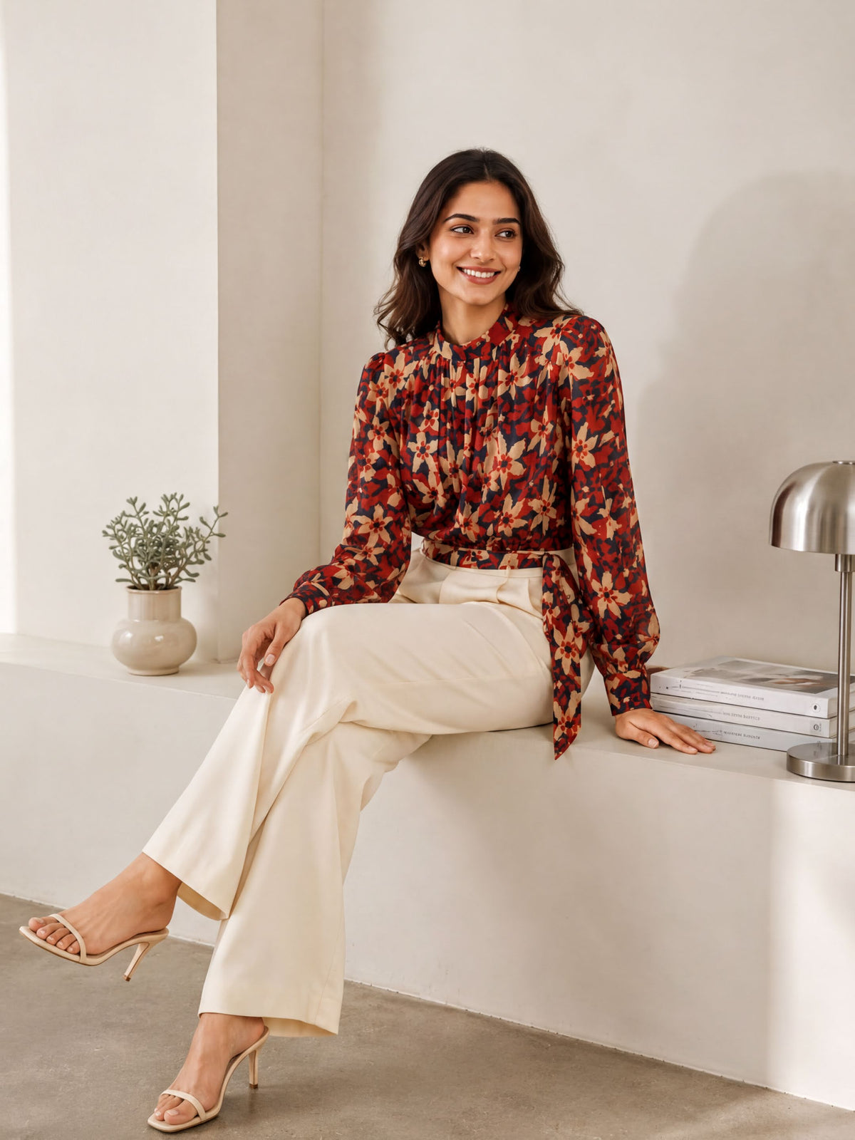 A woman in a floral print blouse and white pants is sitting on a ledge, smiling and looking to the side.