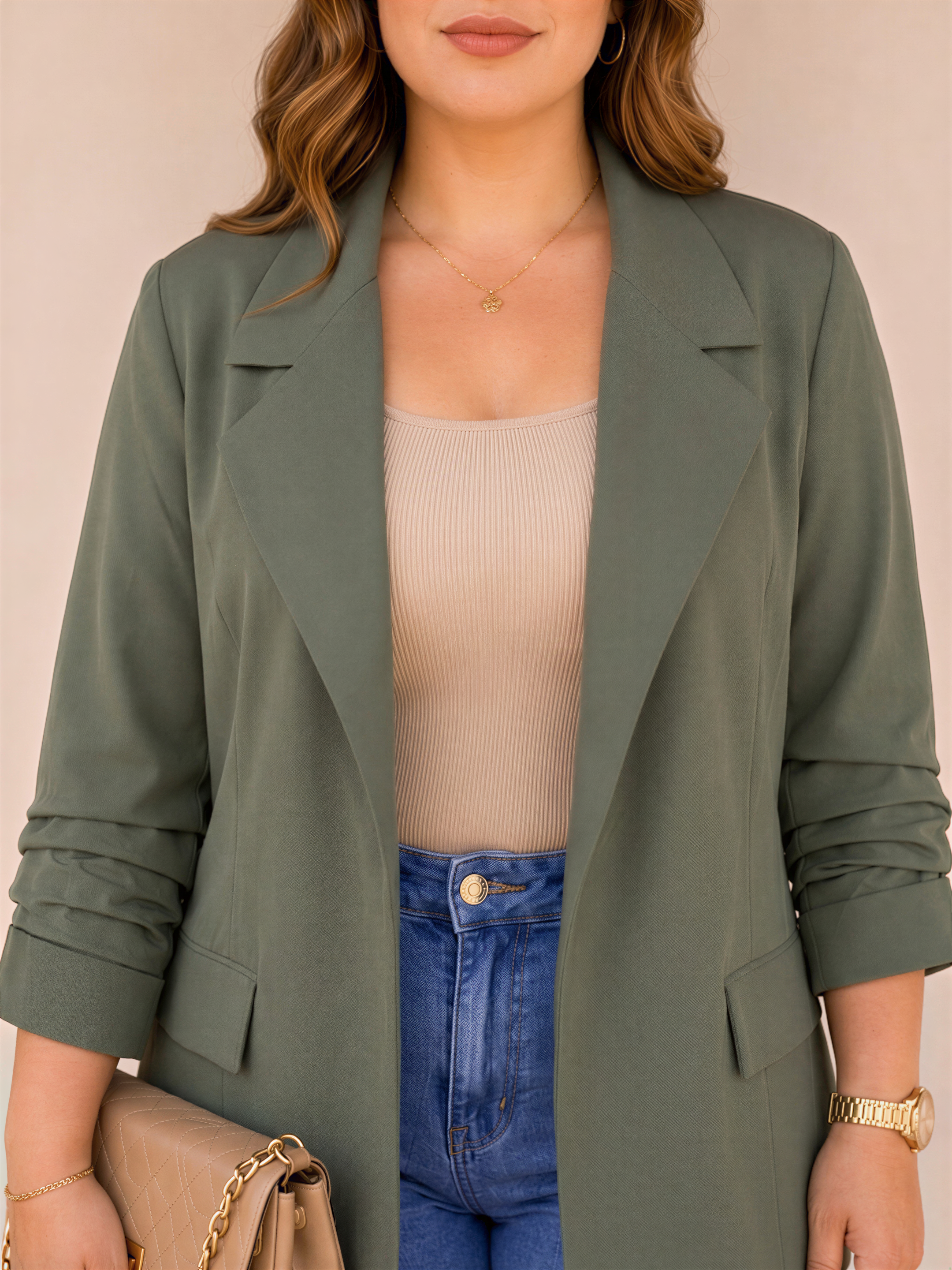 A woman wearing a green blazer, beige top, and blue jeans, holding a beige purse, stands against a light background.