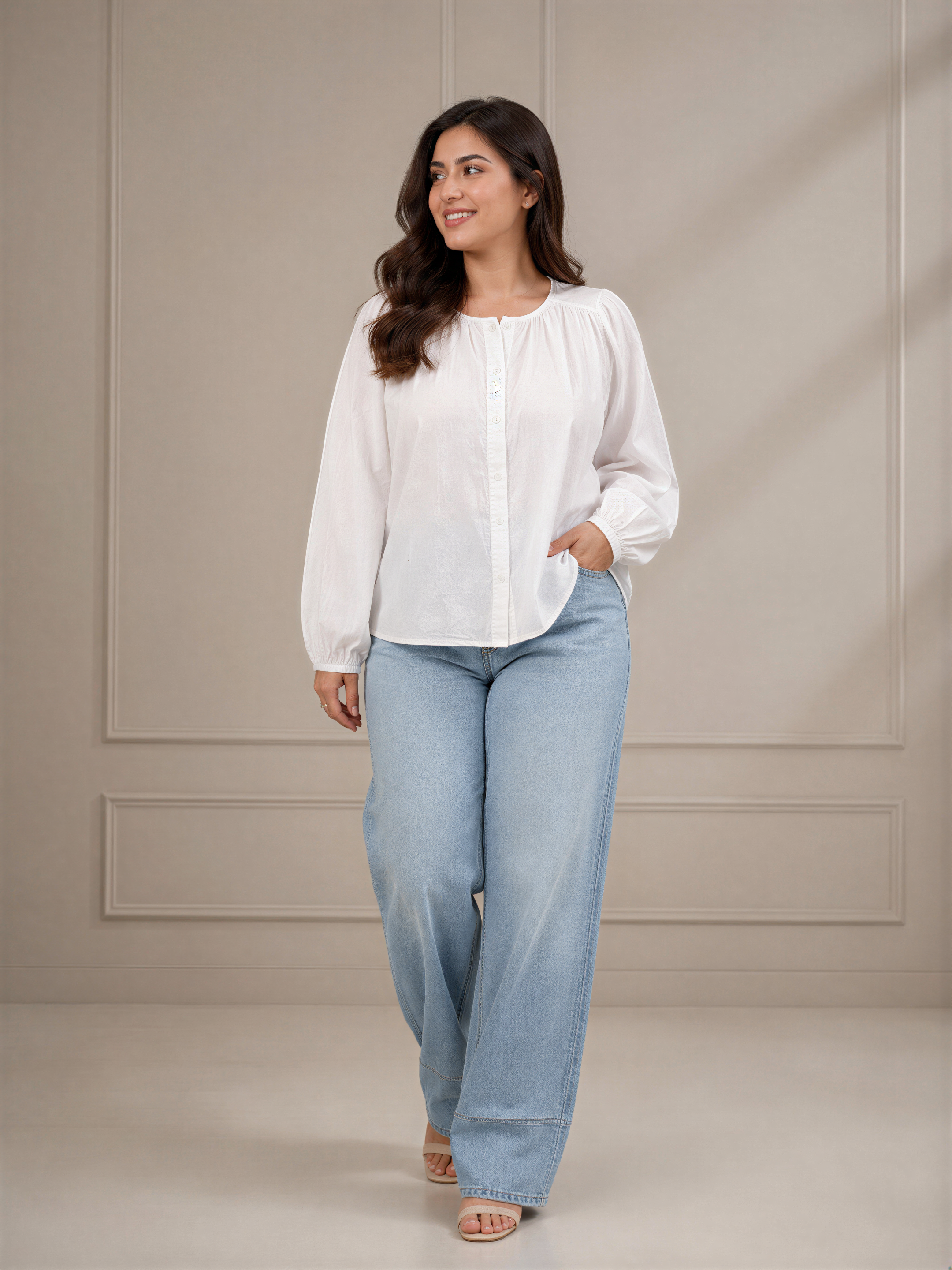 A woman wearing a white blouse and light blue jeans stands in front of a plain beige wall, smiling and looking to her left.