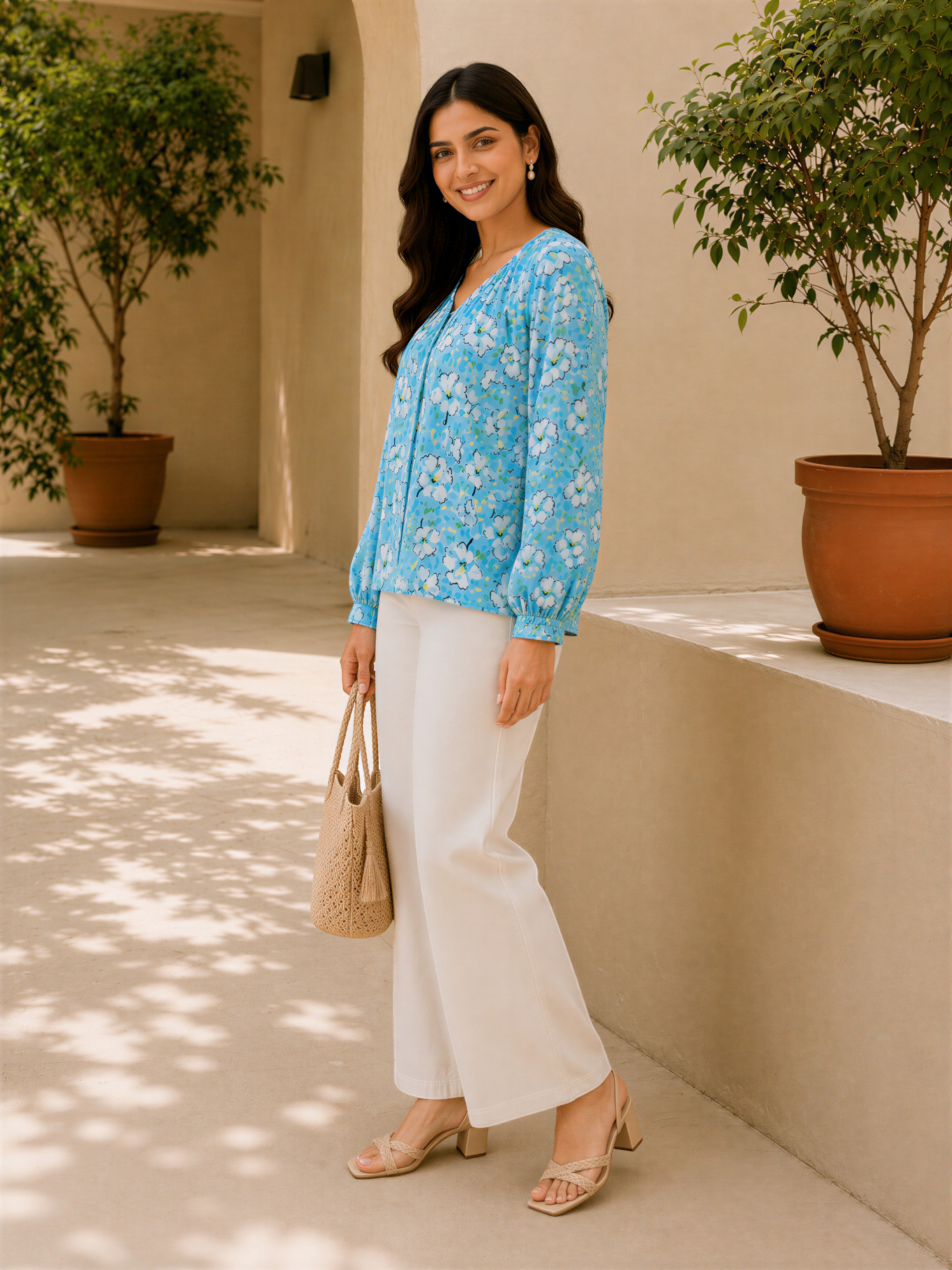 A woman wearing a blue floral blouse and white pants stands in front of a beige wall, holding a beige handbag.