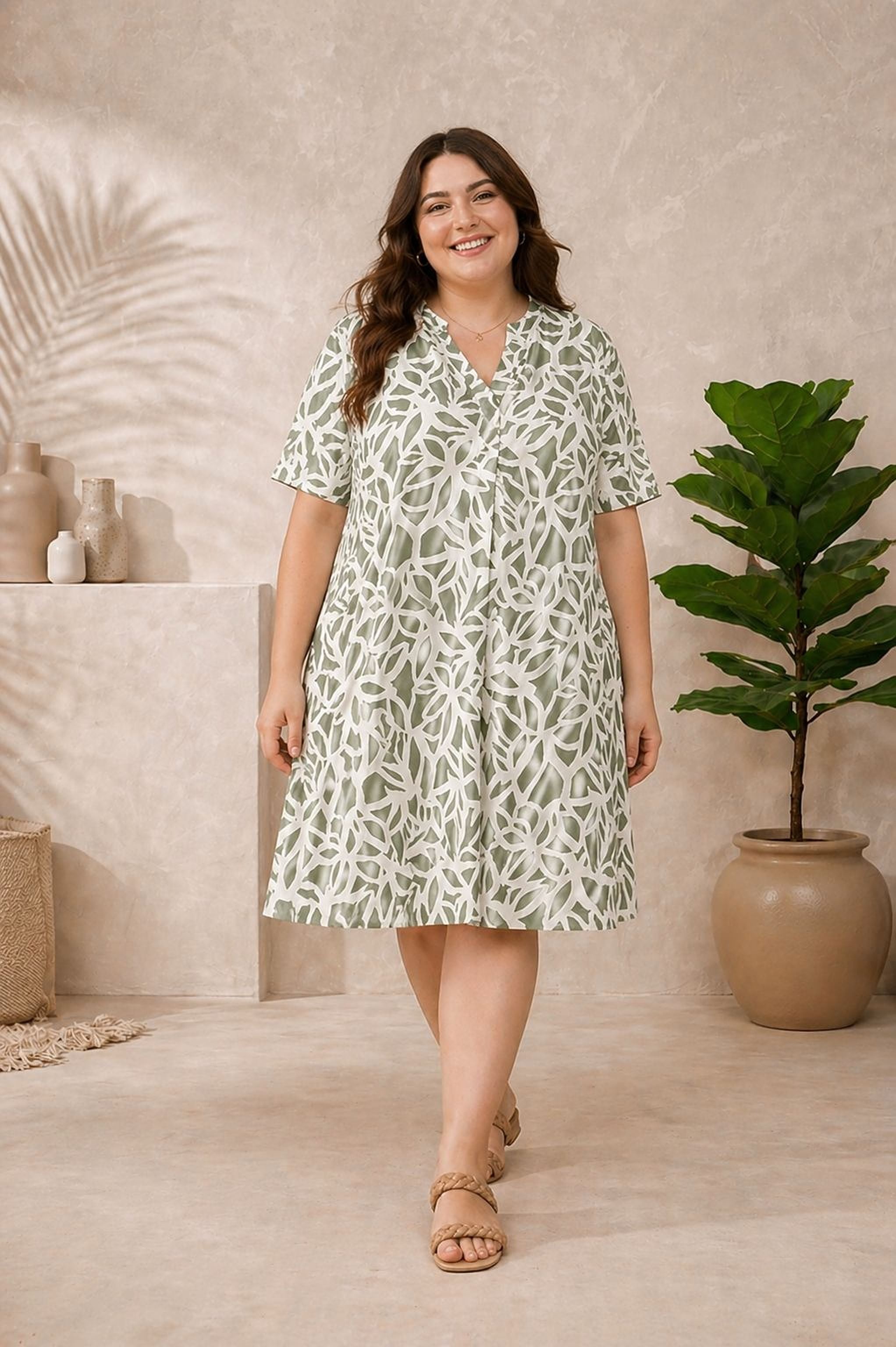 A woman wearing a green and white patterned dress is standing in a room with a potted plant and a shelf with decorative items.