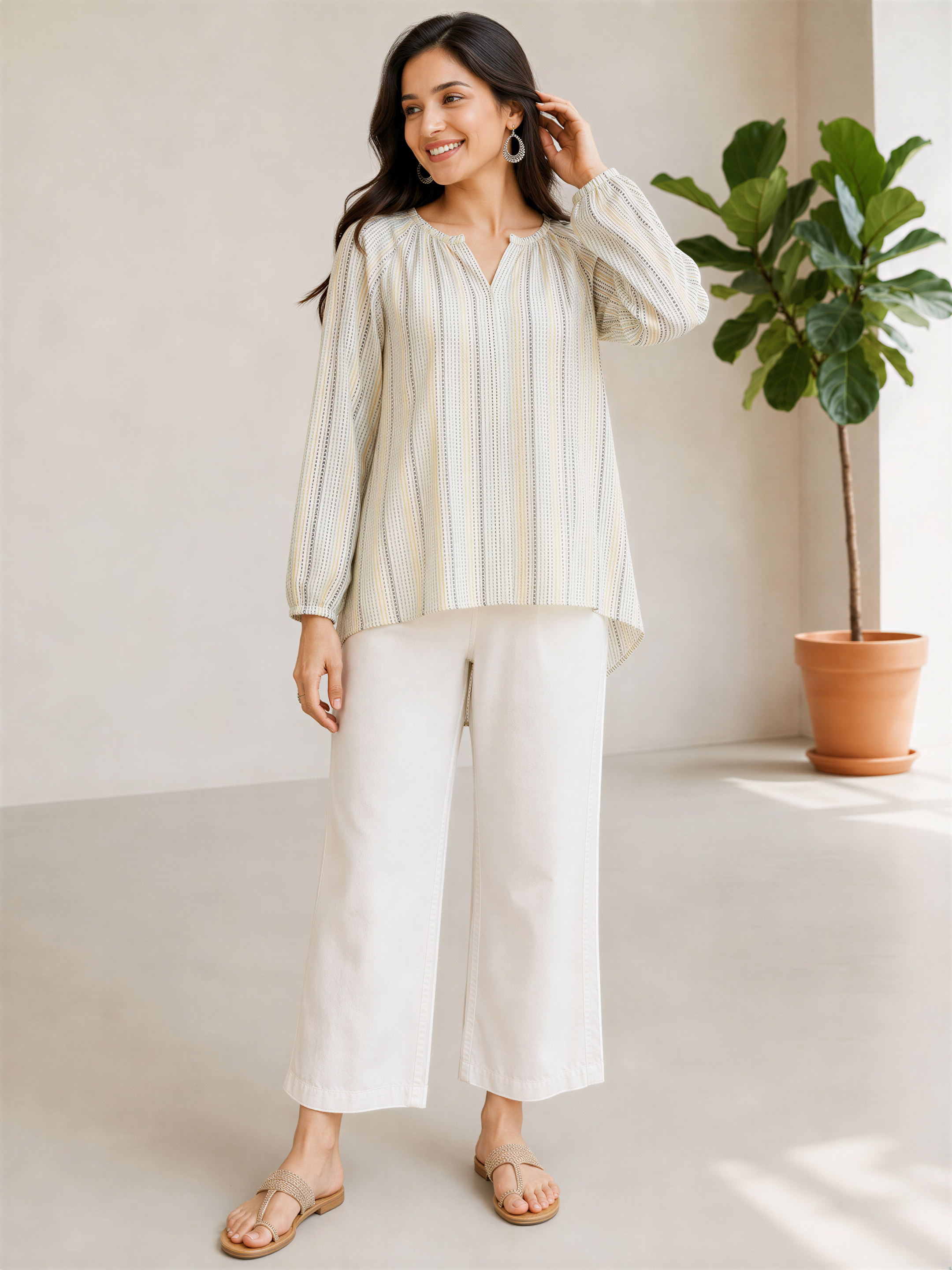 A woman wearing a striped blouse and white pants stands in a room with a potted plant in the background.