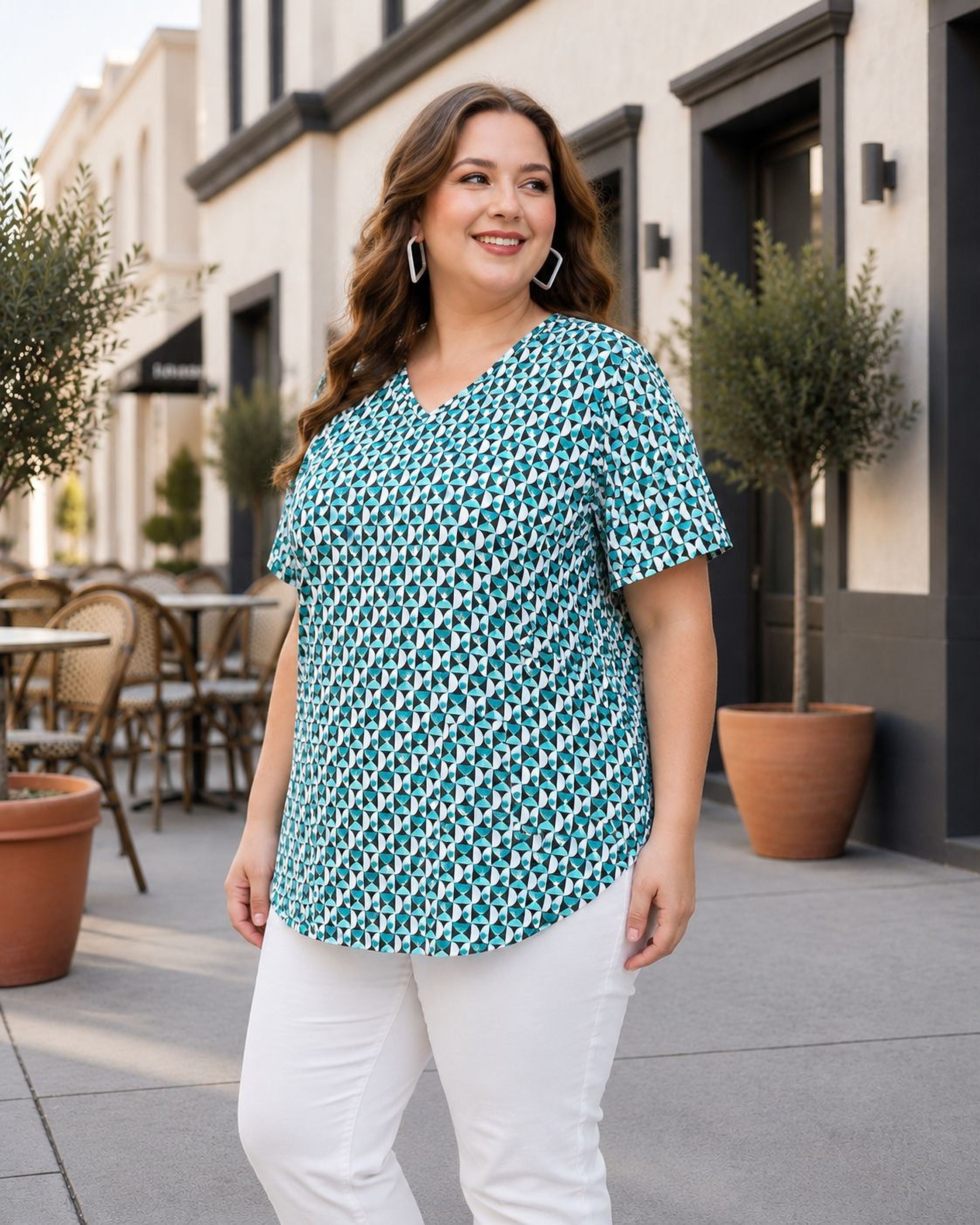 A woman wearing a green and white patterned blouse and white pants is standing on a sidewalk in front of a building with potted plants.