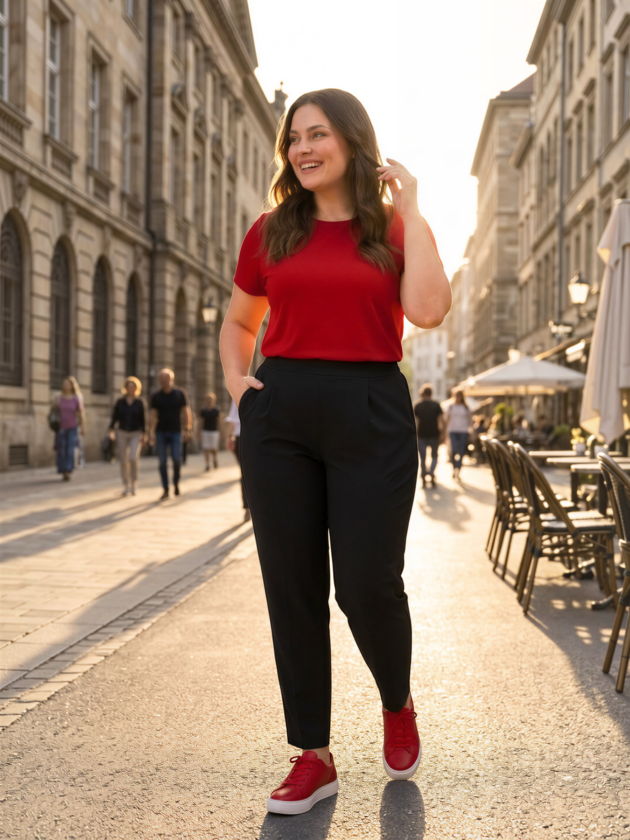 A woman in a red top and black pants walks down a sunlit street, smiling and holding her hand to her face.