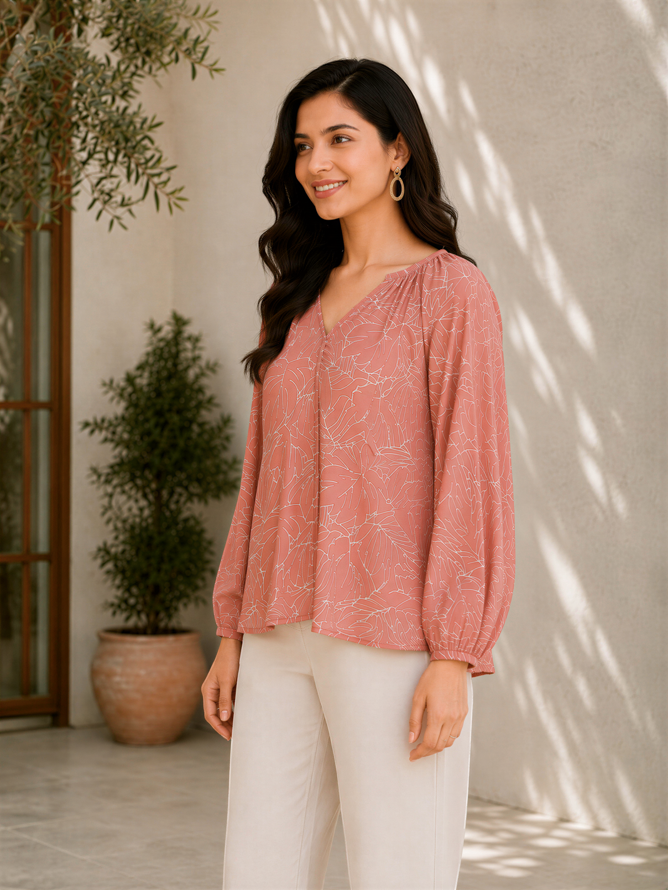 A woman wearing a pink blouse with a floral pattern and white pants stands in front of a white wall with a potted plant.