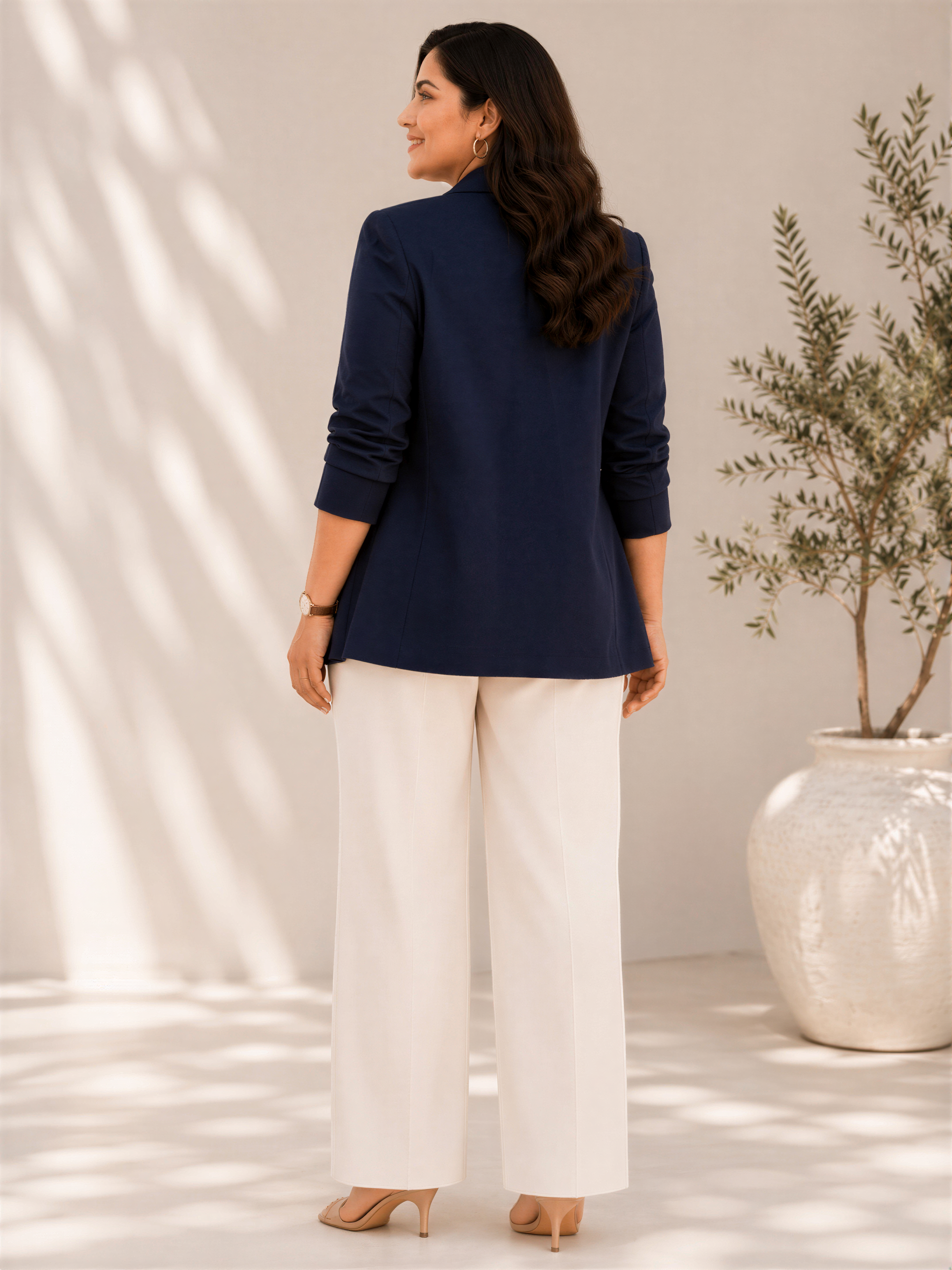 A woman in a navy blue blazer and white pants stands in front of a white vase with greenery, smiling at the camera.