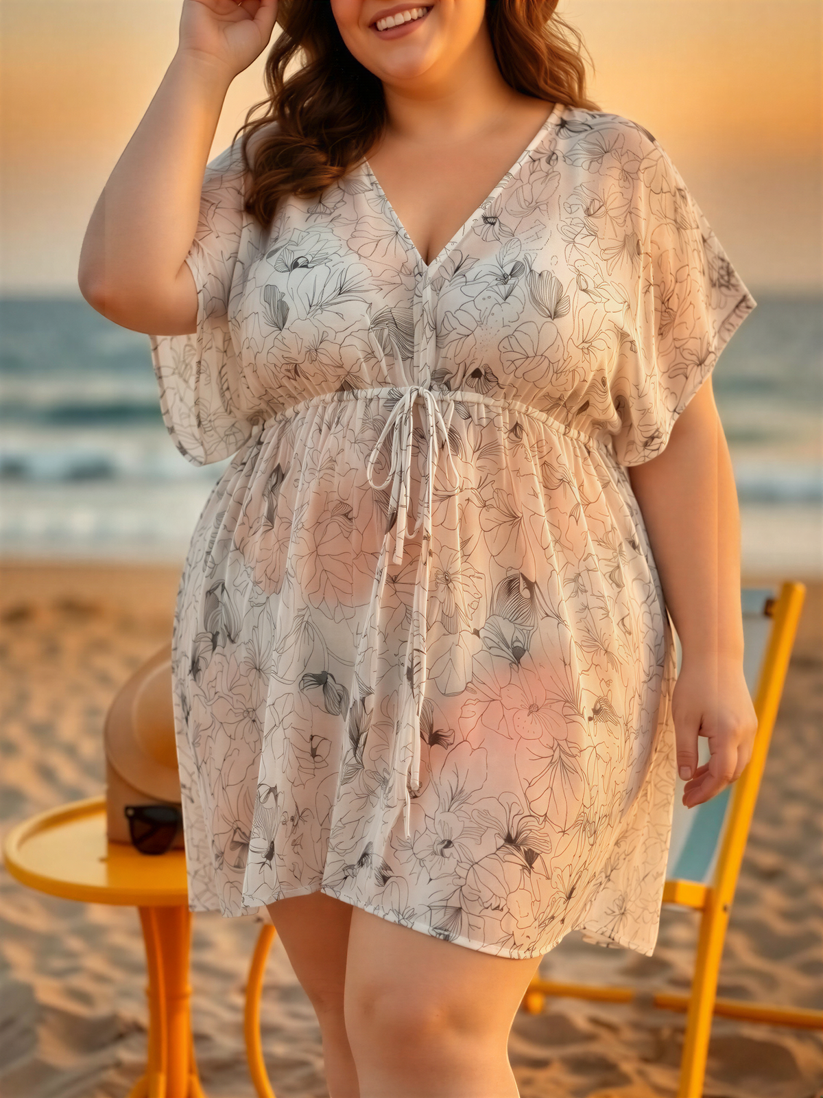 A woman wearing a floral print dress stands on a beach, smiling and looking towards the camera.
