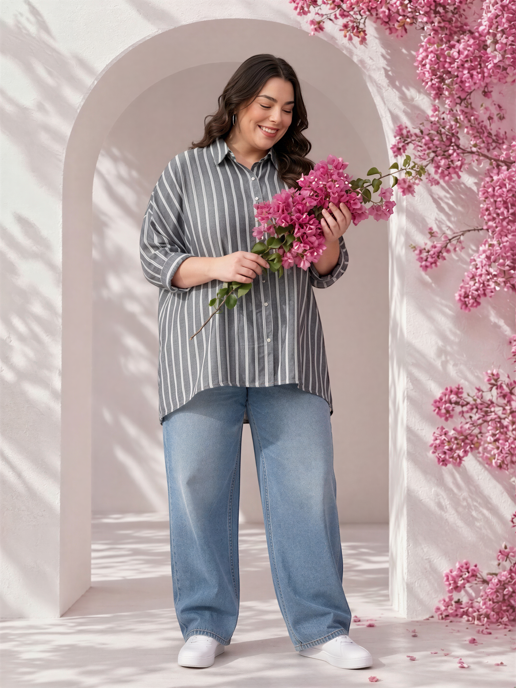 A woman in a striped shirt and jeans stands in front of a pink flowering tree, holding a bouquet of pink flowers.