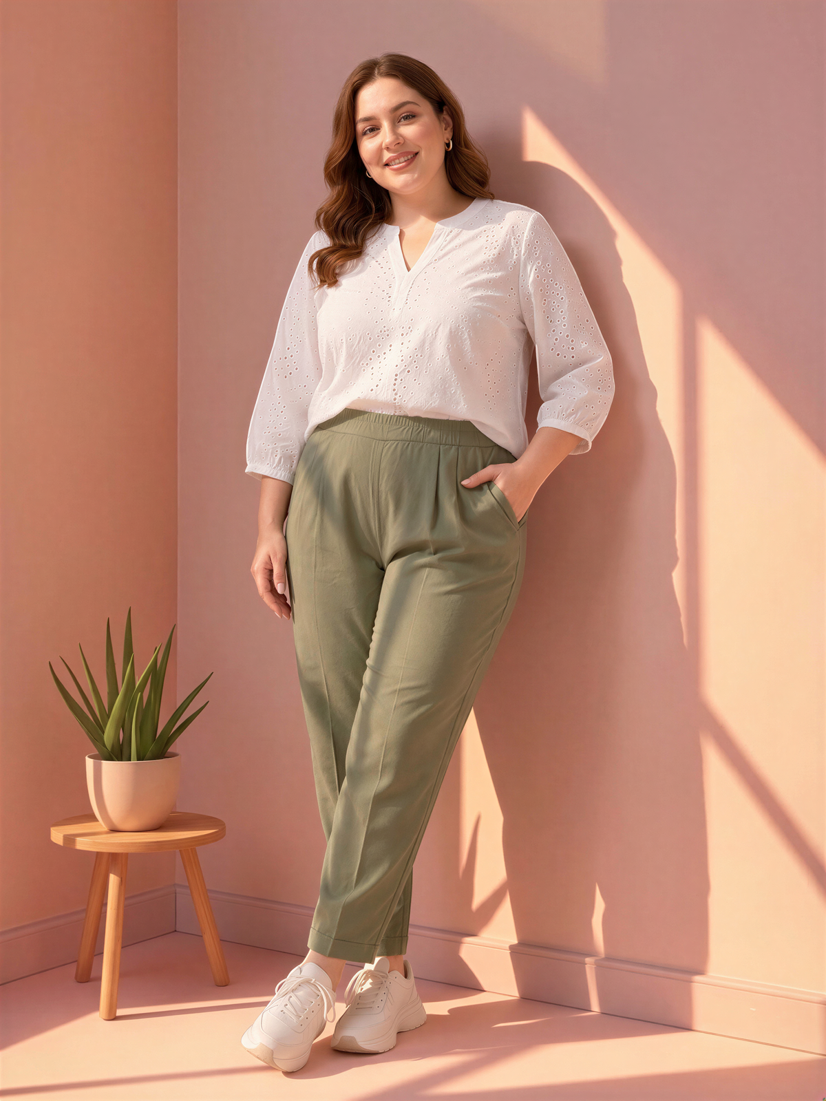 A woman in a white blouse and olive green pants stands in a room with a pink wall, smiling and leaning against the wall.