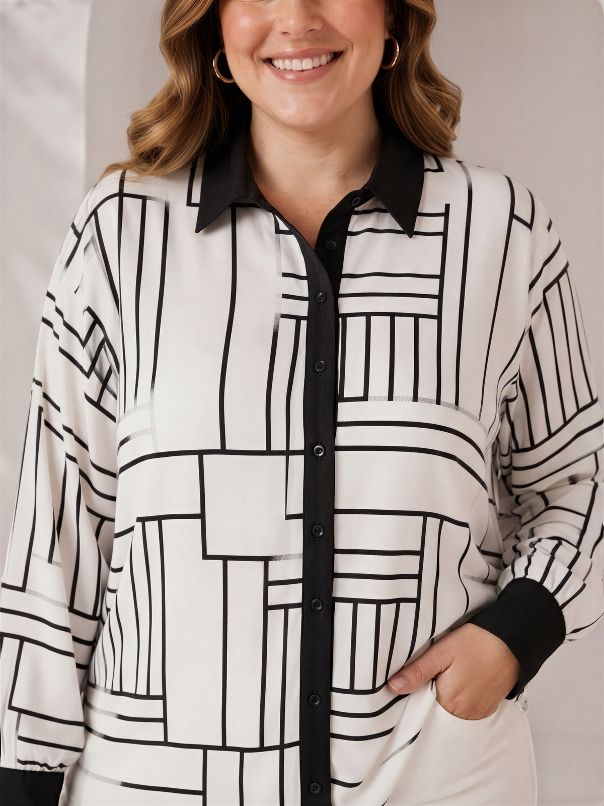 A woman with long, wavy brown hair is smiling and wearing a white and black patterned blouse with a black collar.