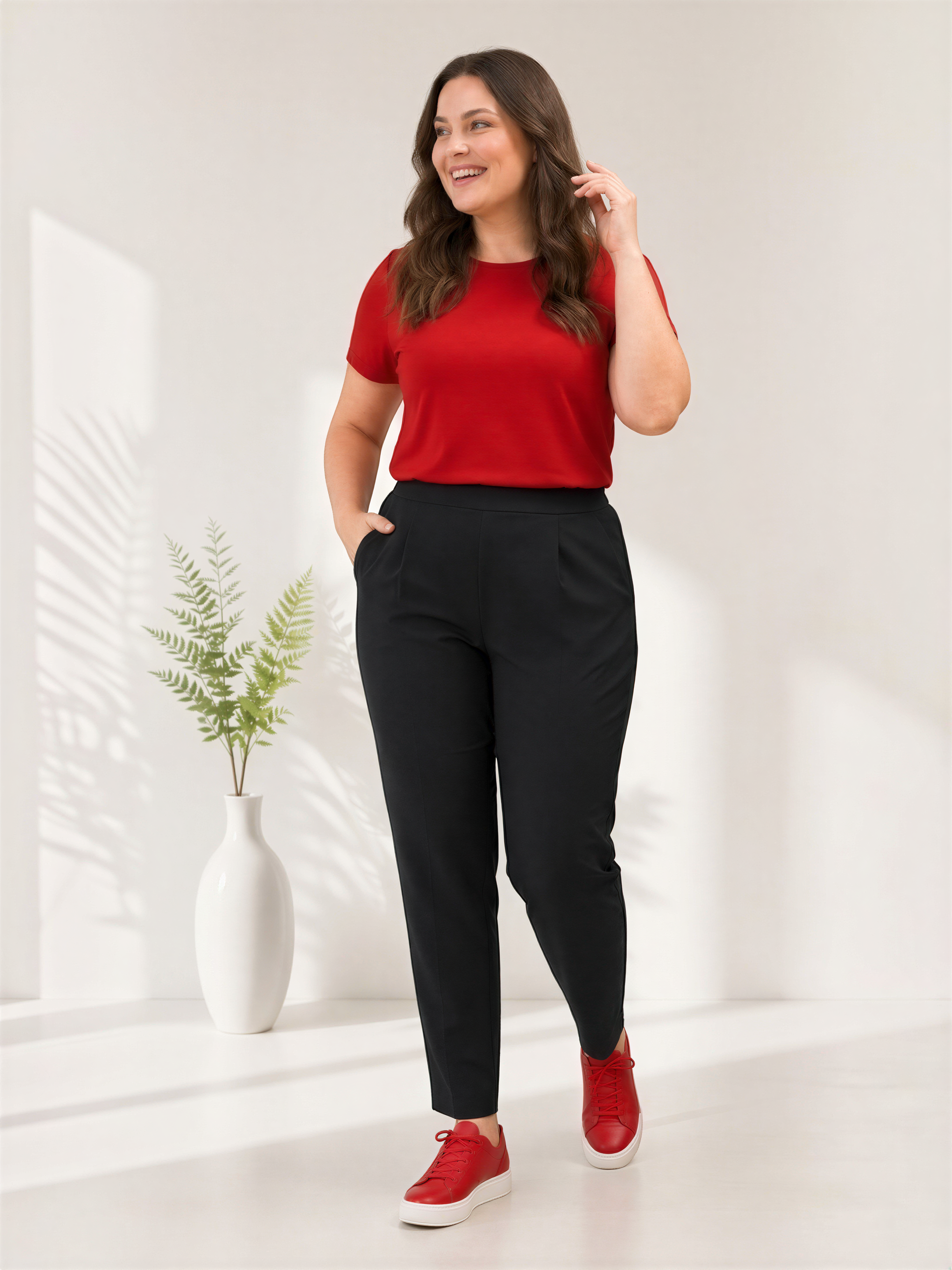 A woman in a red top and black pants stands in a room with a white vase of ferns on a shelf to her left.