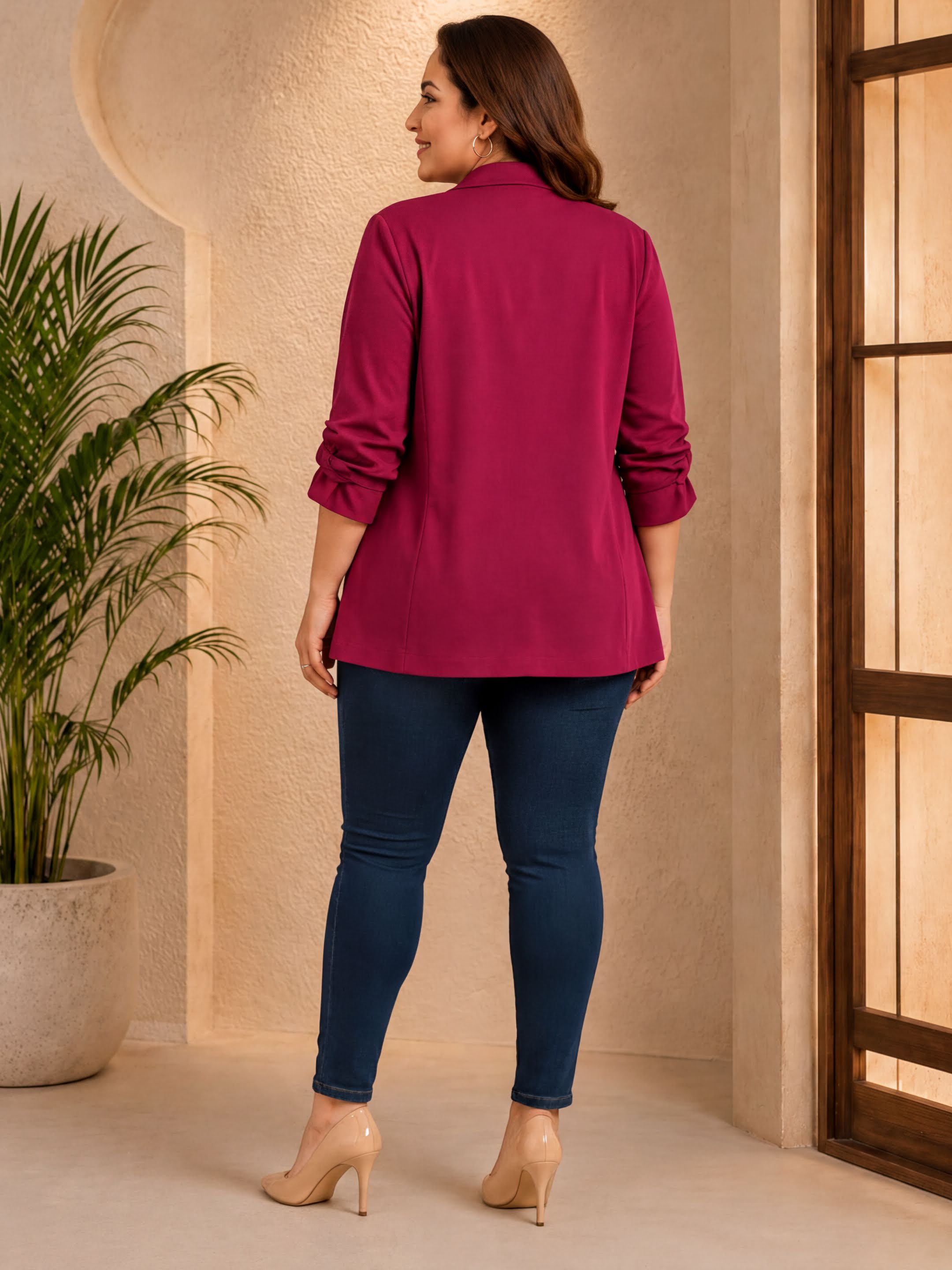 A woman wearing a red blazer and blue jeans stands in a room with a potted plant and a window.