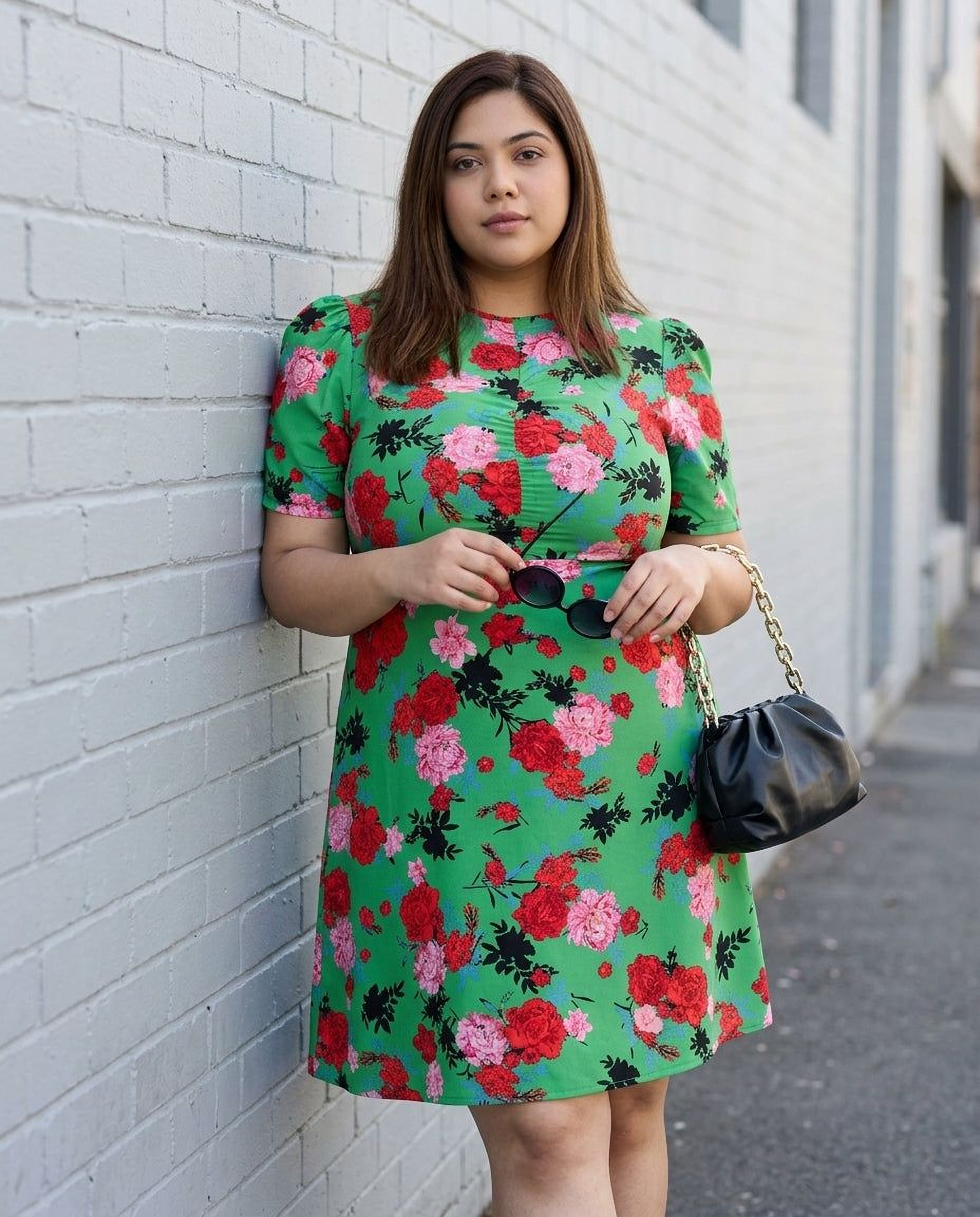 A woman wearing a green floral print dress and sunglasses, holding a black purse, and smiling at the camera.