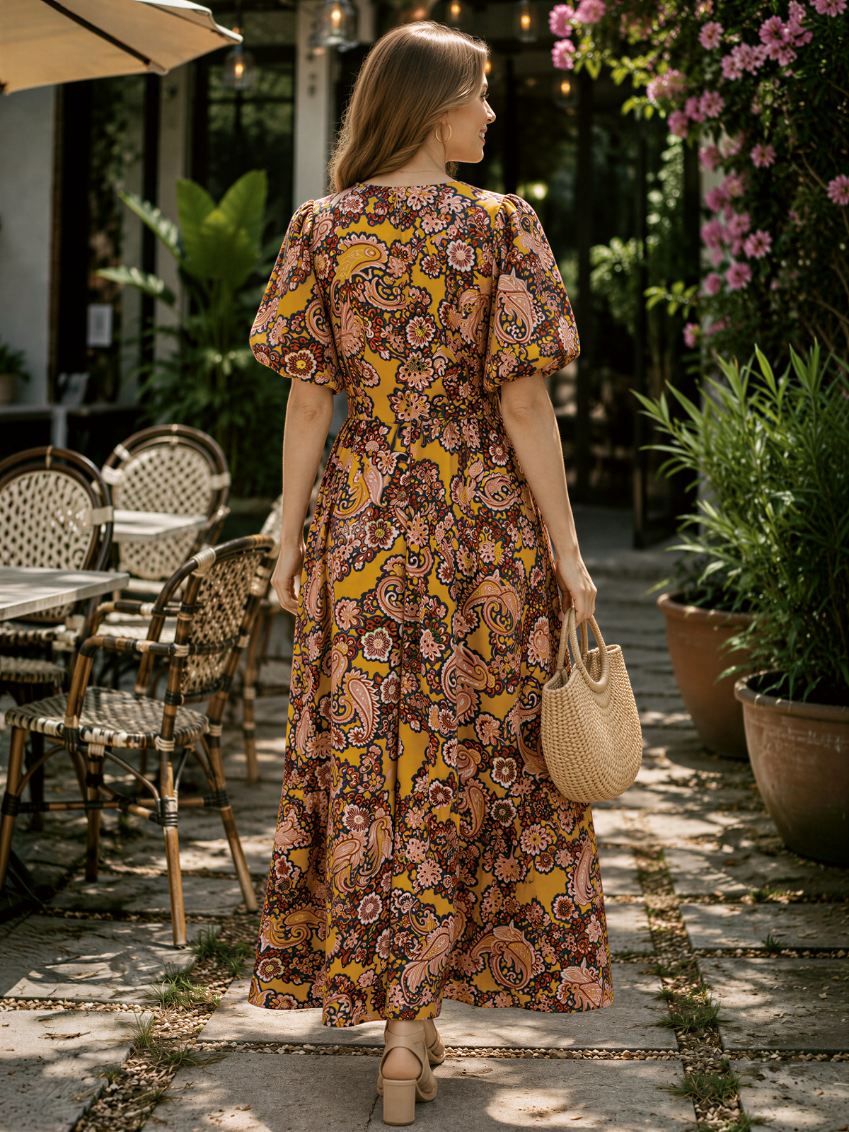 A woman wearing a long, colorful dress with a paisley pattern walks down a path lined with potted plants and outdoor furniture.