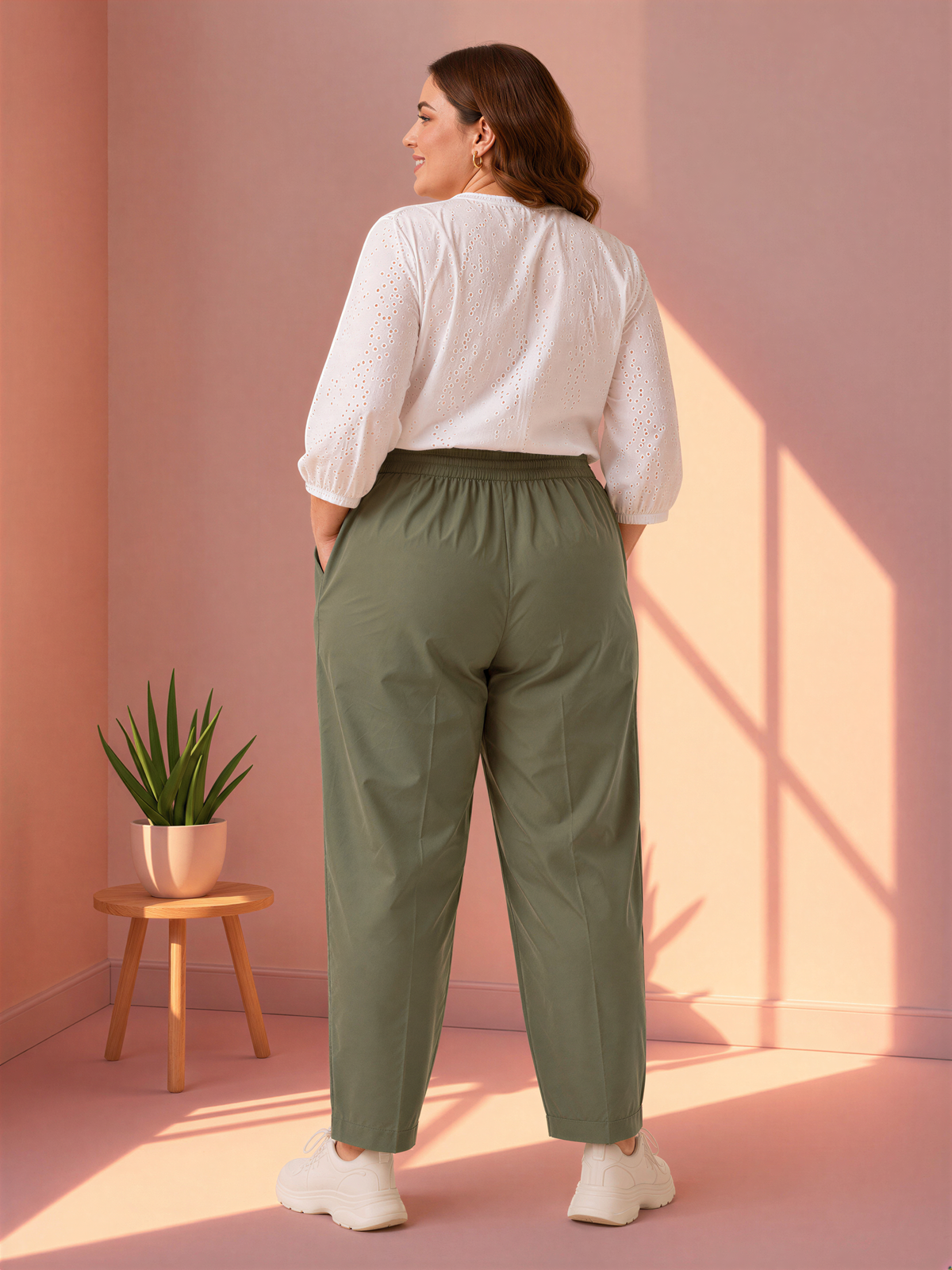 A woman wearing a white blouse and olive green pants stands in a room with a pink wall, looking away from the camera.