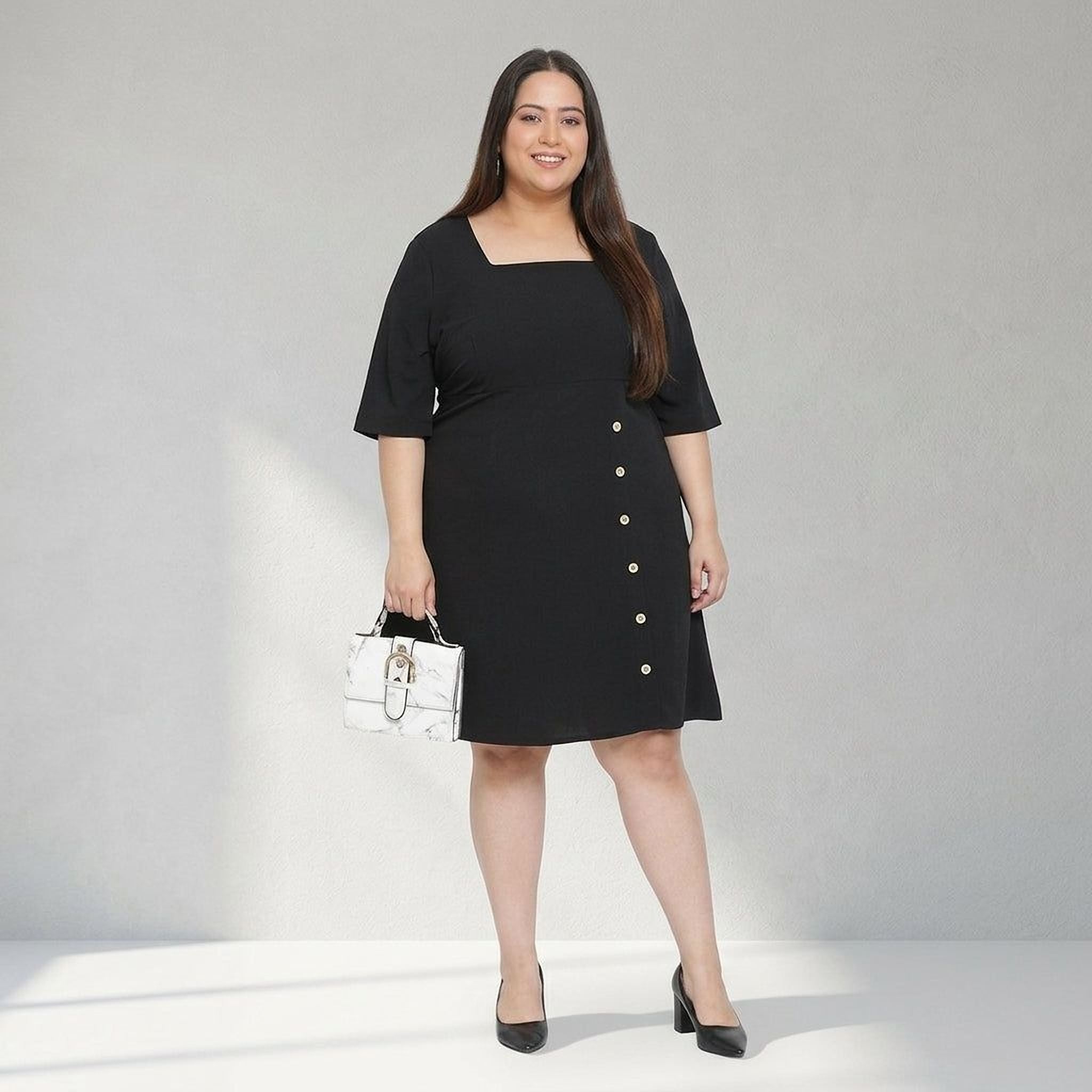 Plus size woman in black square neck dress with button detail, holding handbag, standing indoors