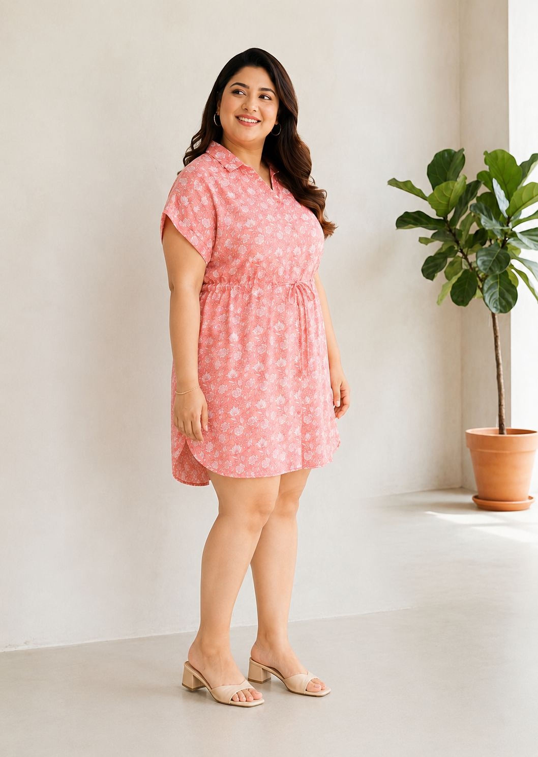 A woman wearing a pink floral dress stands in a room with a potted plant in the background.