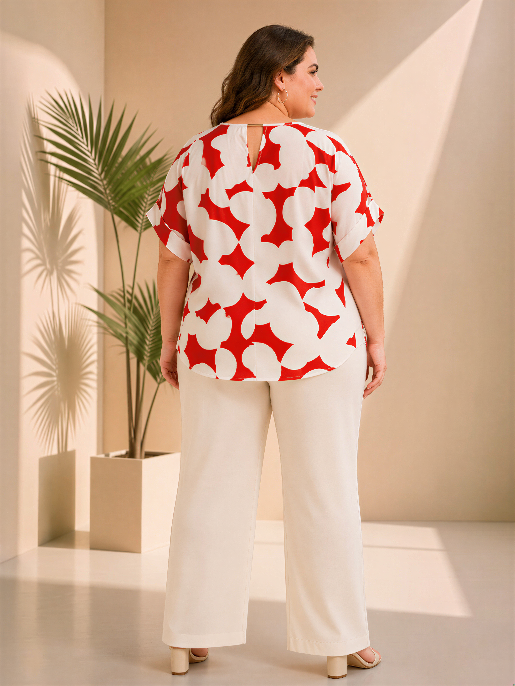 A woman wearing a red and white patterned blouse and white pants stands in a room with a potted plant in the background.