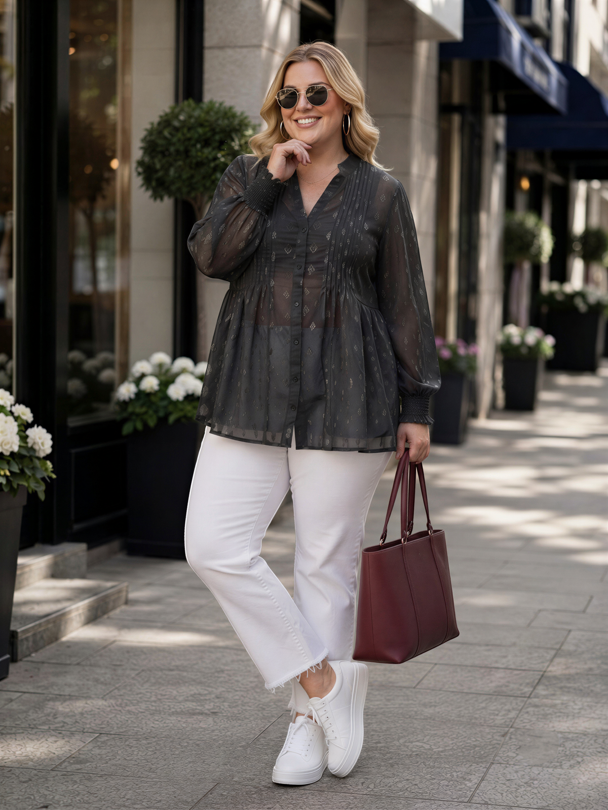 A woman wearing a black and gray blouse, white pants, and white sneakers is standing on a sidewalk, smiling and holding a red handbag.