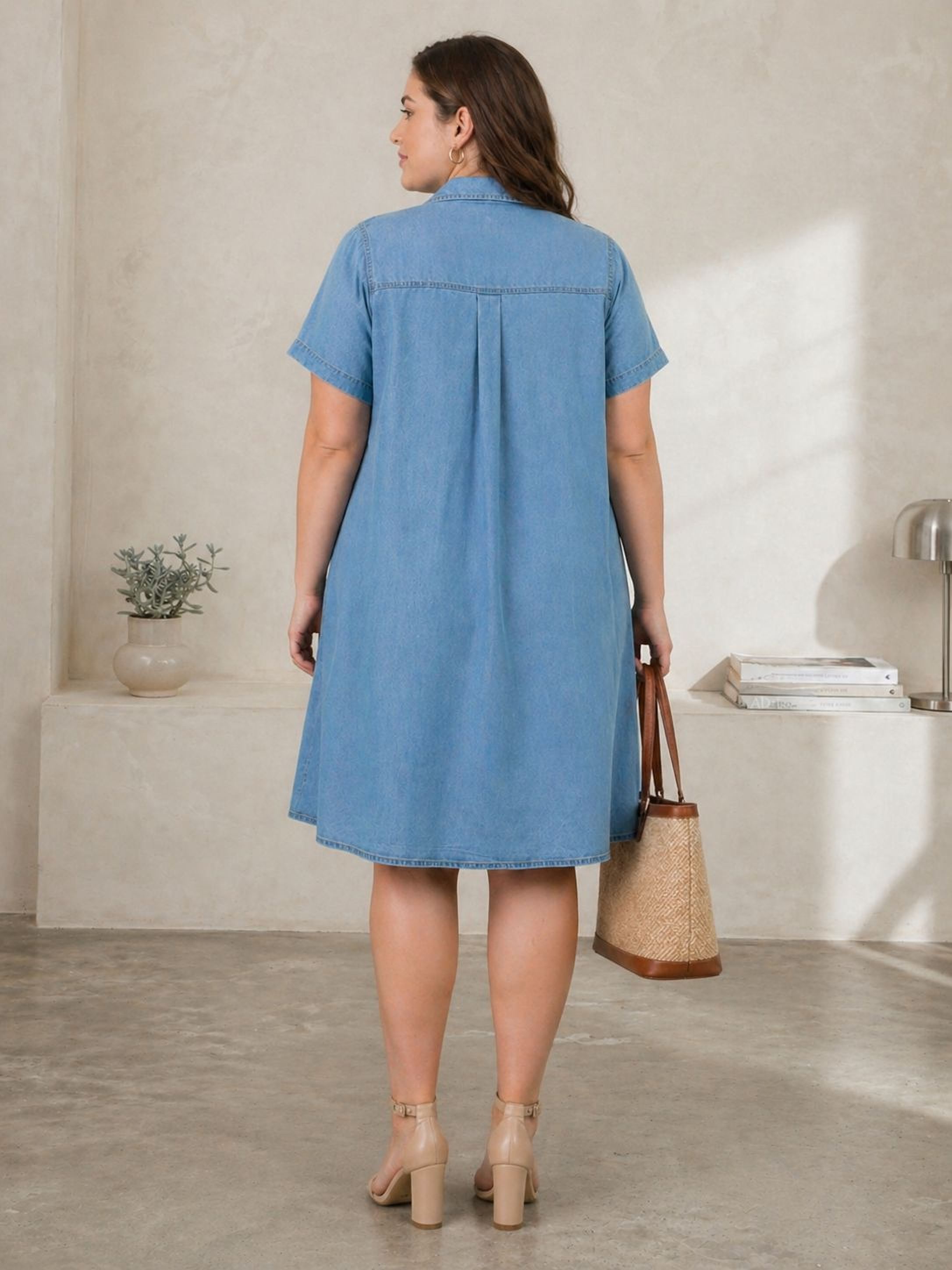 A woman wearing a blue denim dress and carrying a woven handbag stands in a room with a minimalist design.