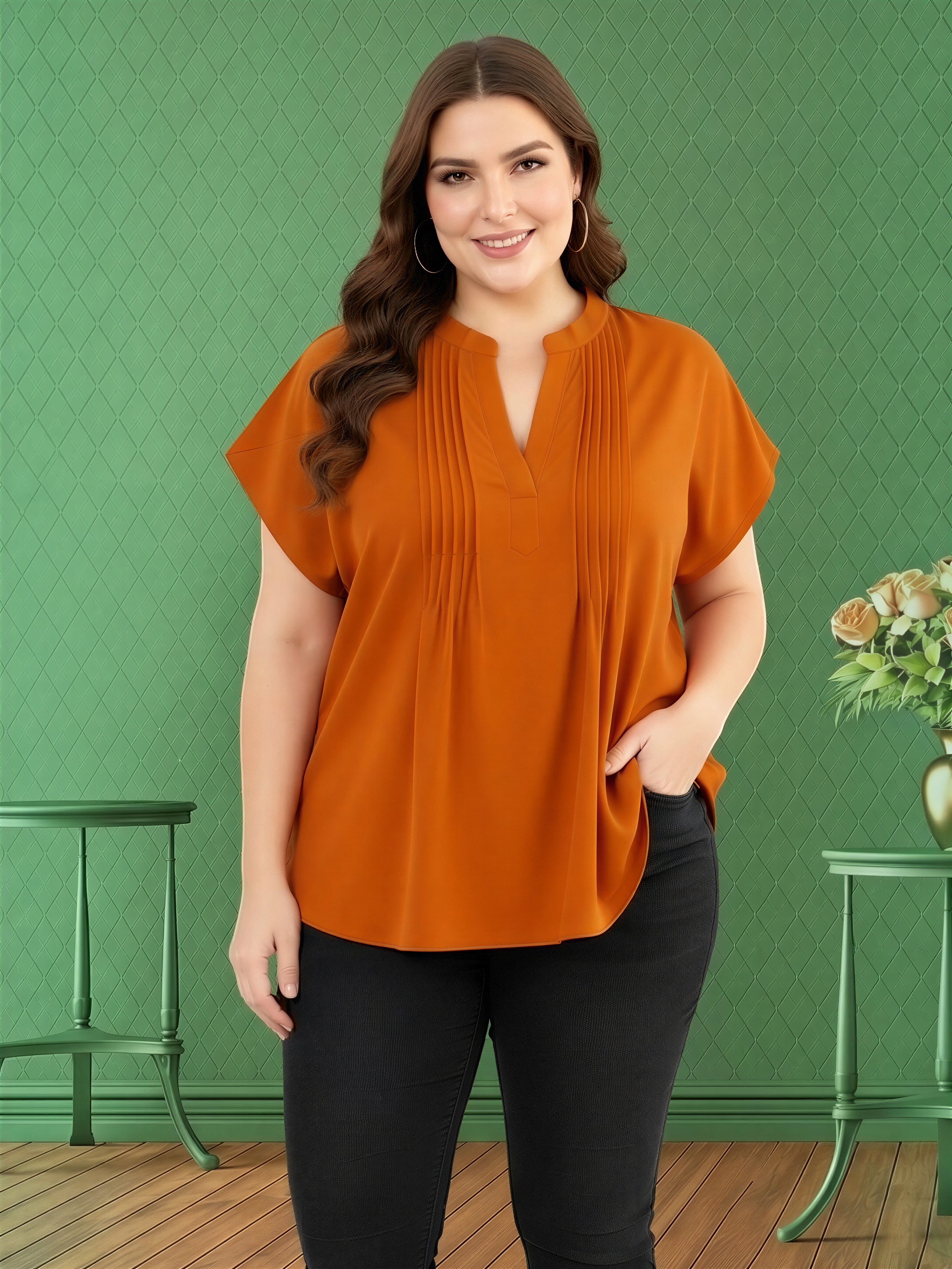 A woman with long brown hair is standing in front of a green wall, wearing an orange blouse and black pants.