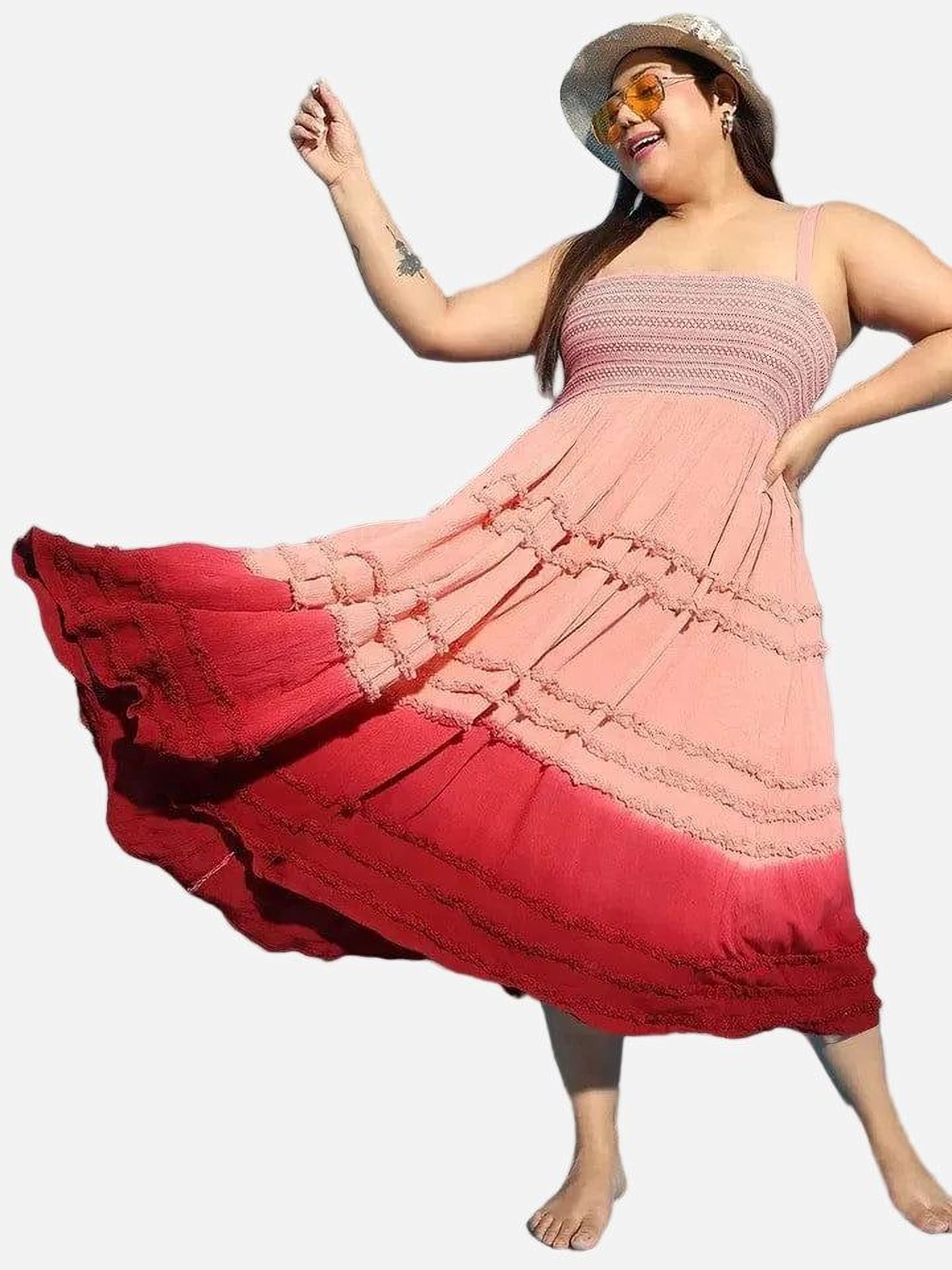 A woman wearing a pink and red tiered dress and a wide-brimmed hat is smiling and gesturing with her hand.
