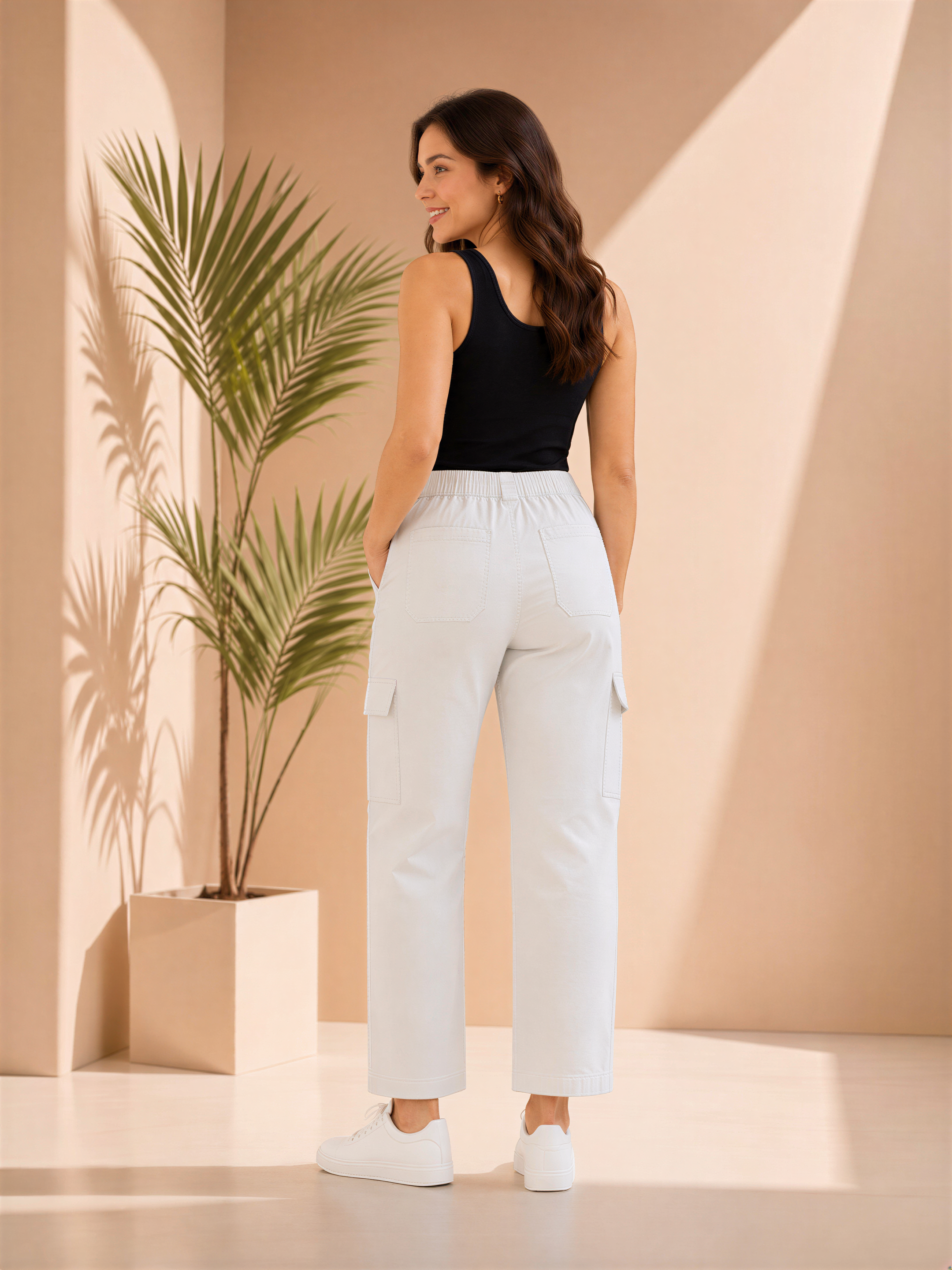 A woman in a black top and white pants stands in a room with a potted plant, facing away from the camera.