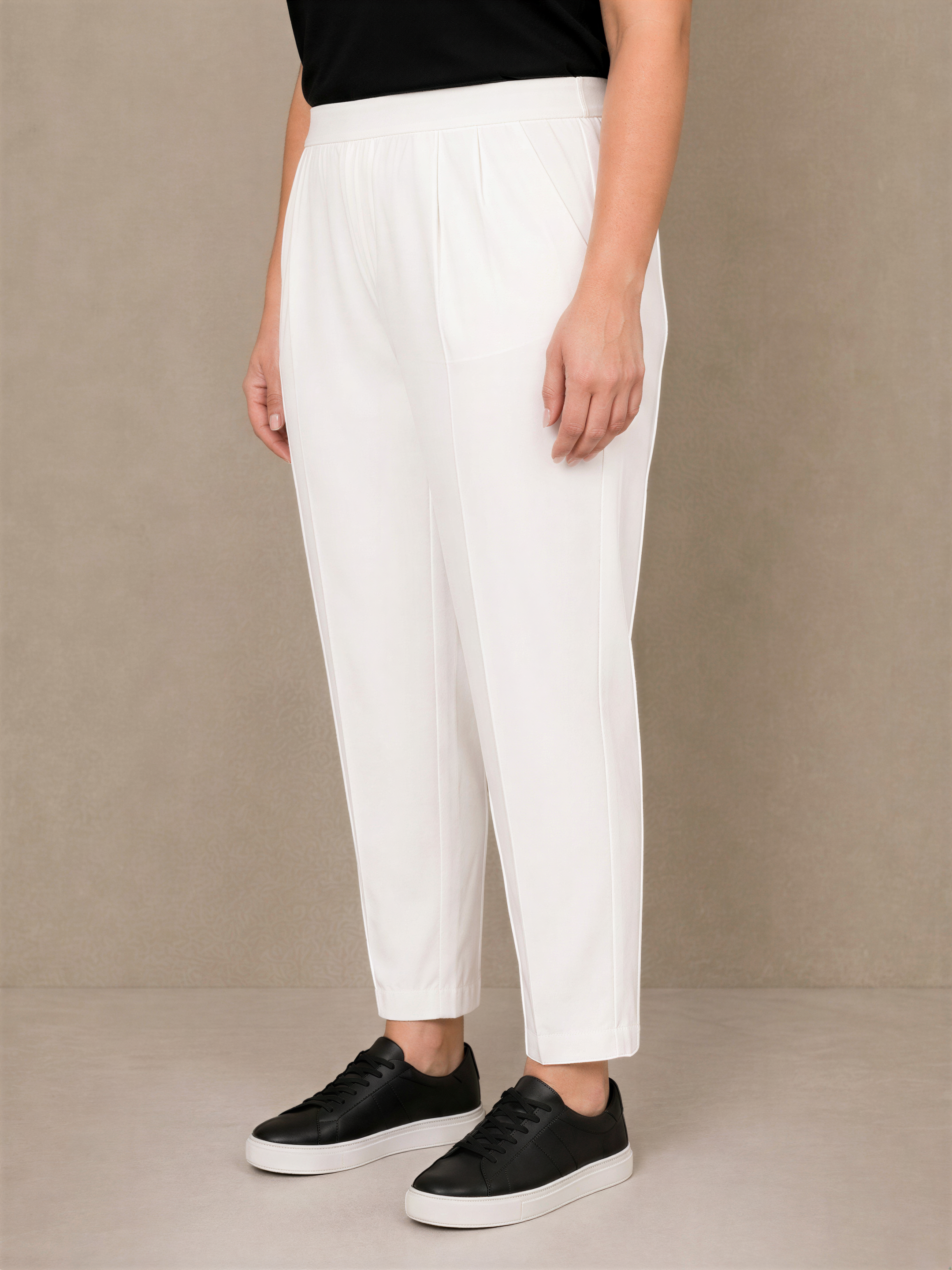 A person wearing white pants and black shoes stands against a beige background.