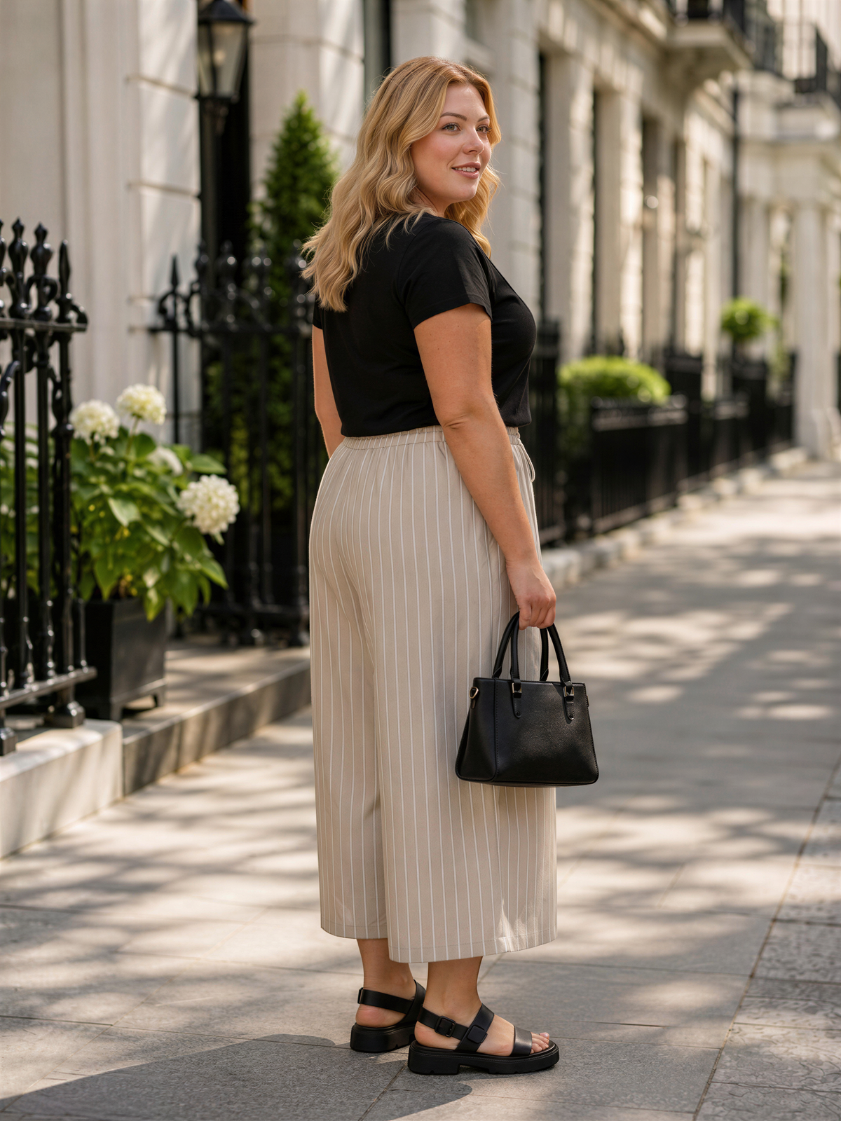 A woman in a black top and beige striped skirt stands on a sidewalk, holding a black handbag and smiling.