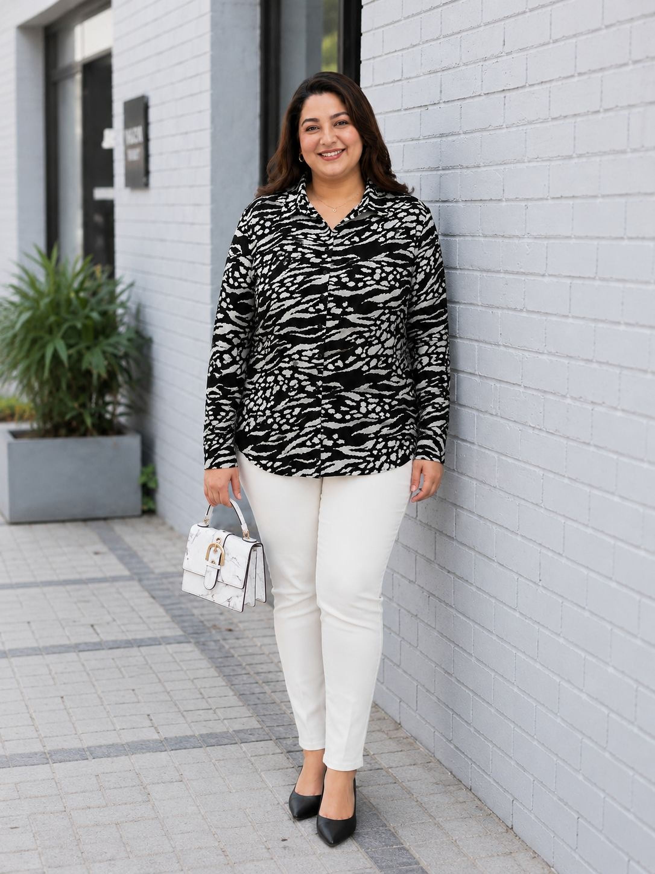 A woman wearing a black and white patterned blouse and white pants is standing in front of a white brick wall, smiling and holding a white handbag.