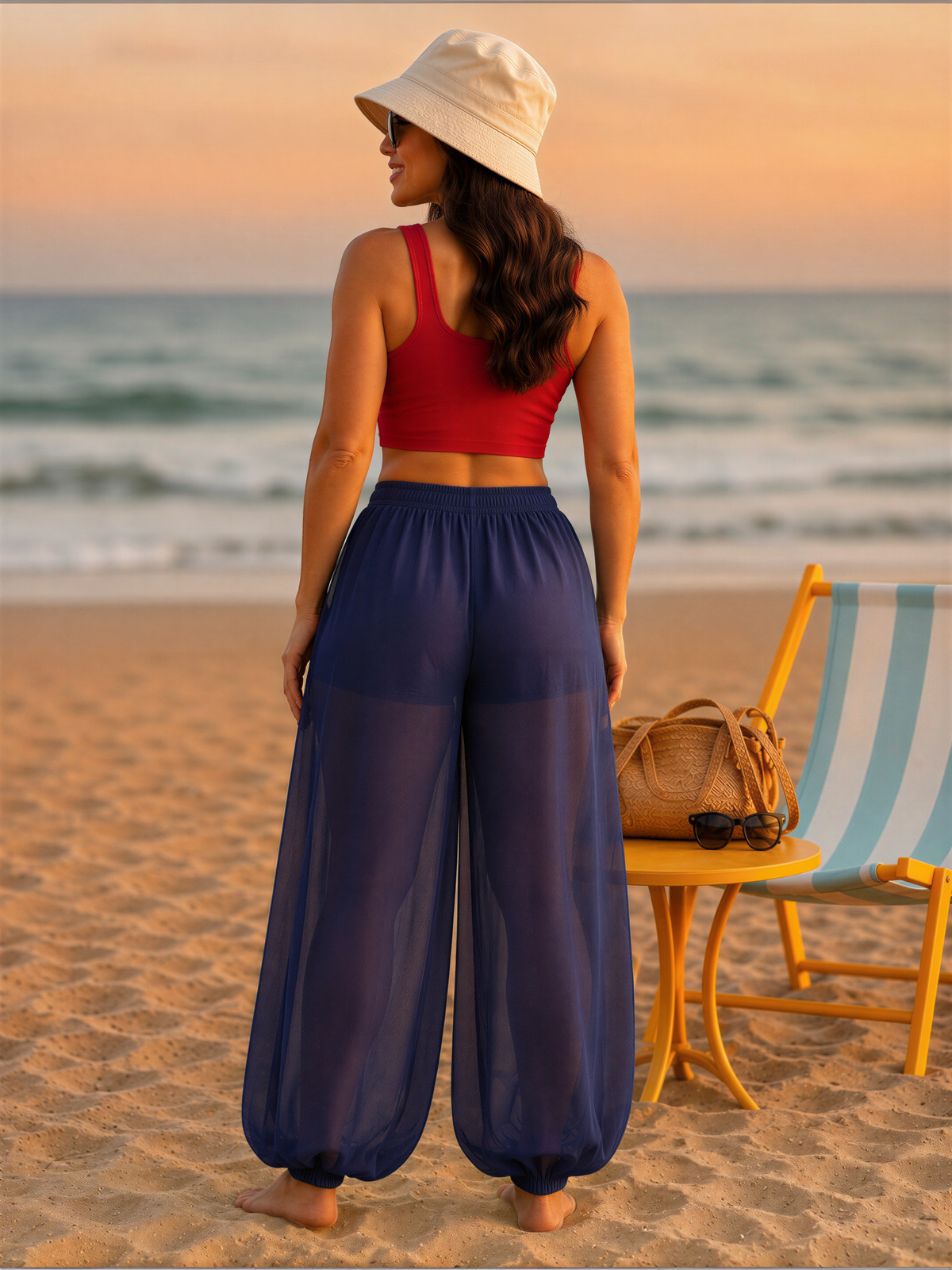 A woman in a red top and blue pants stands on a beach, facing the ocean, with a yellow chair and a handbag nearby.