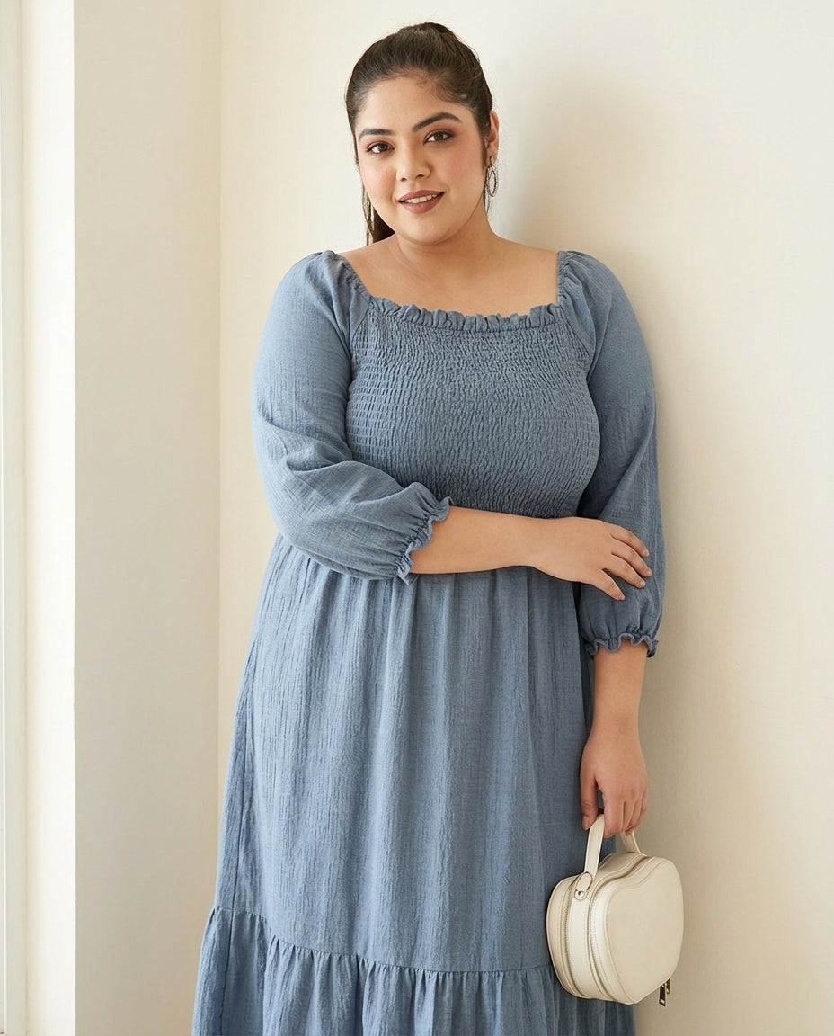 Plus size woman in a blue smocked dress holding a small white handbag, standing by a wall.