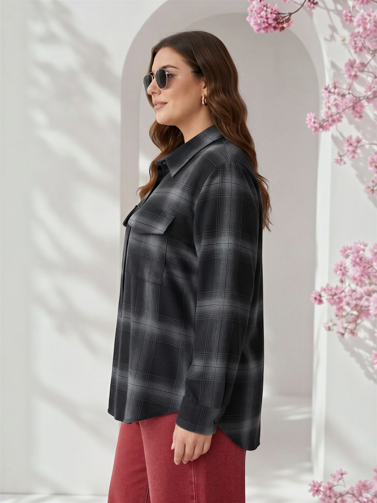 A woman wearing a black and gray plaid shirt and sunglasses stands in front of a white archway with pink flowers.