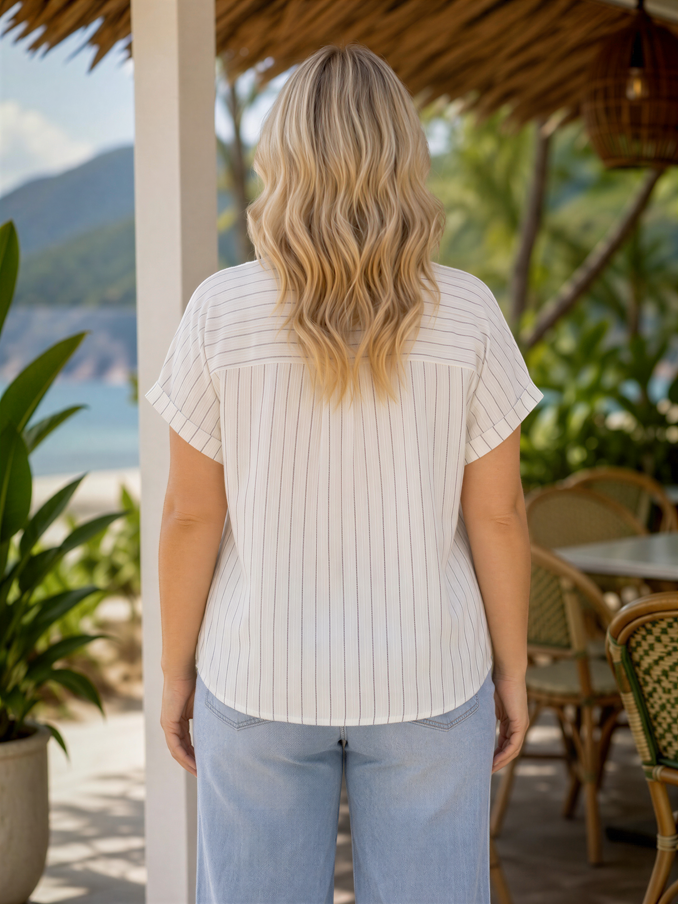 A woman with long, wavy blonde hair is standing in front of a tropical beach setting, with palm trees and a thatched roof structure visible in the background.