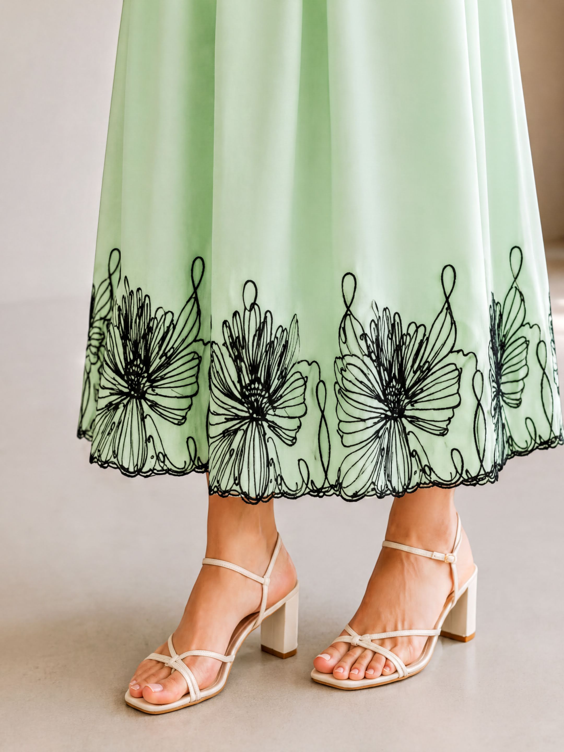 A woman wearing a mint green dress with black floral embroidery on the skirt and strappy white strappy heels.