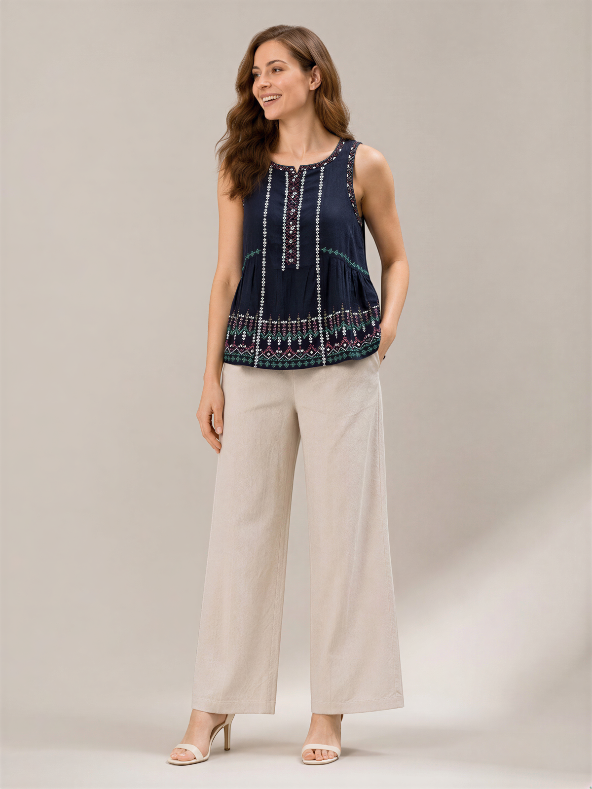 A woman wearing a navy blue sleeveless top with a patterned design, beige wide-leg pants, and high heels, smiling and posing for the camera.