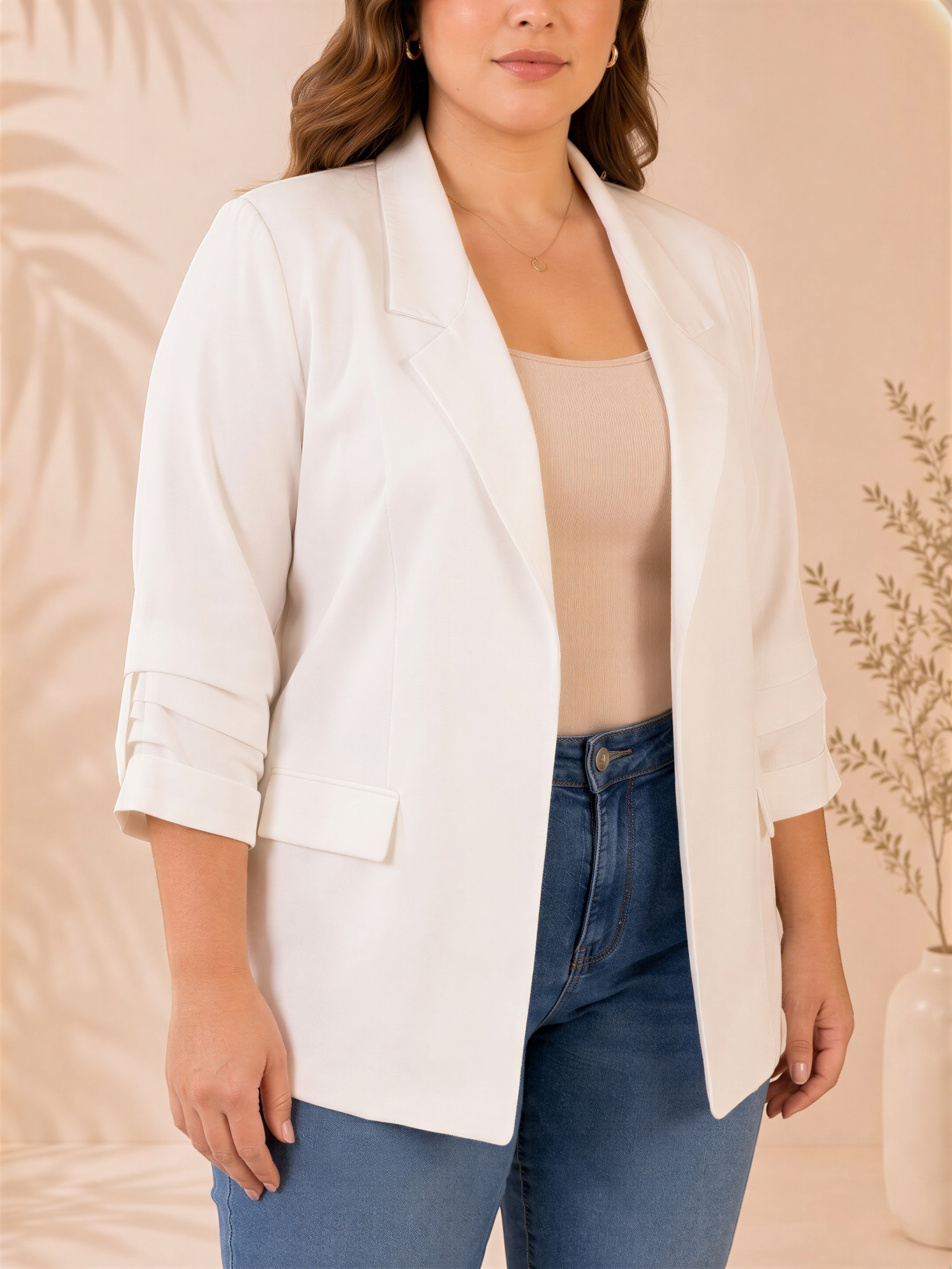 A woman wearing a white blazer and blue jeans stands in front of a pink background with a plant.