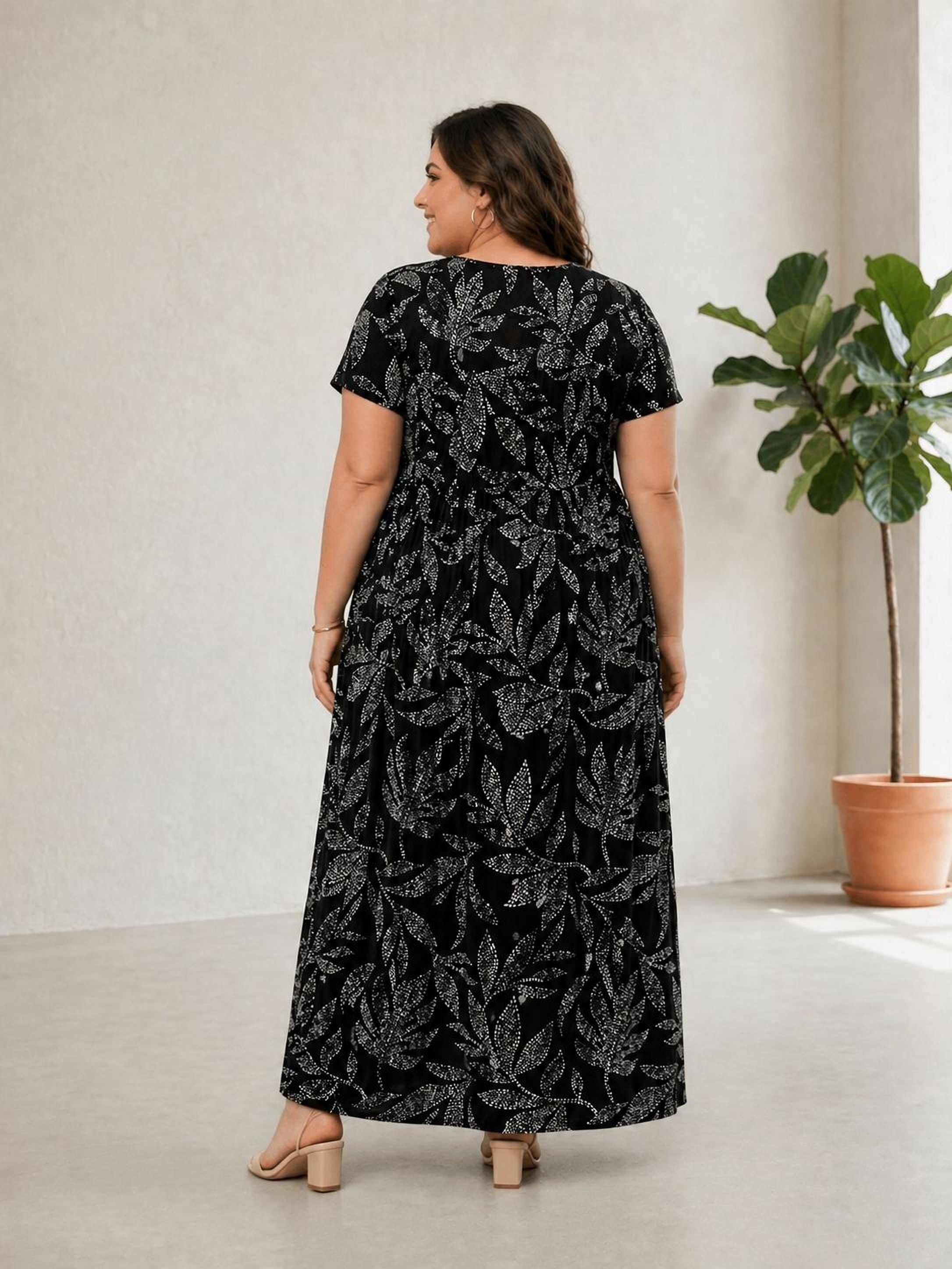 A woman wearing a long black dress with a leaf pattern stands in a room with a potted plant.