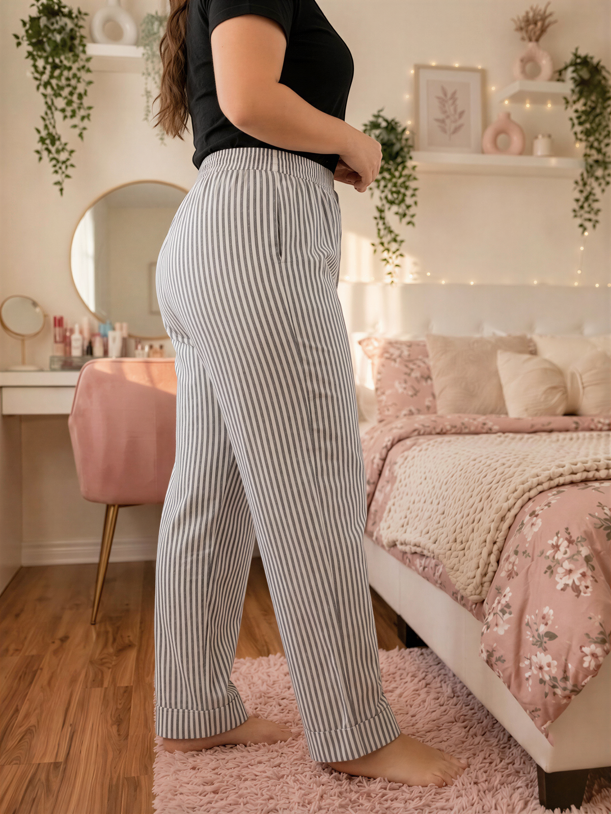 A person wearing striped pants stands in a bedroom with a bed, plants, and a vanity.