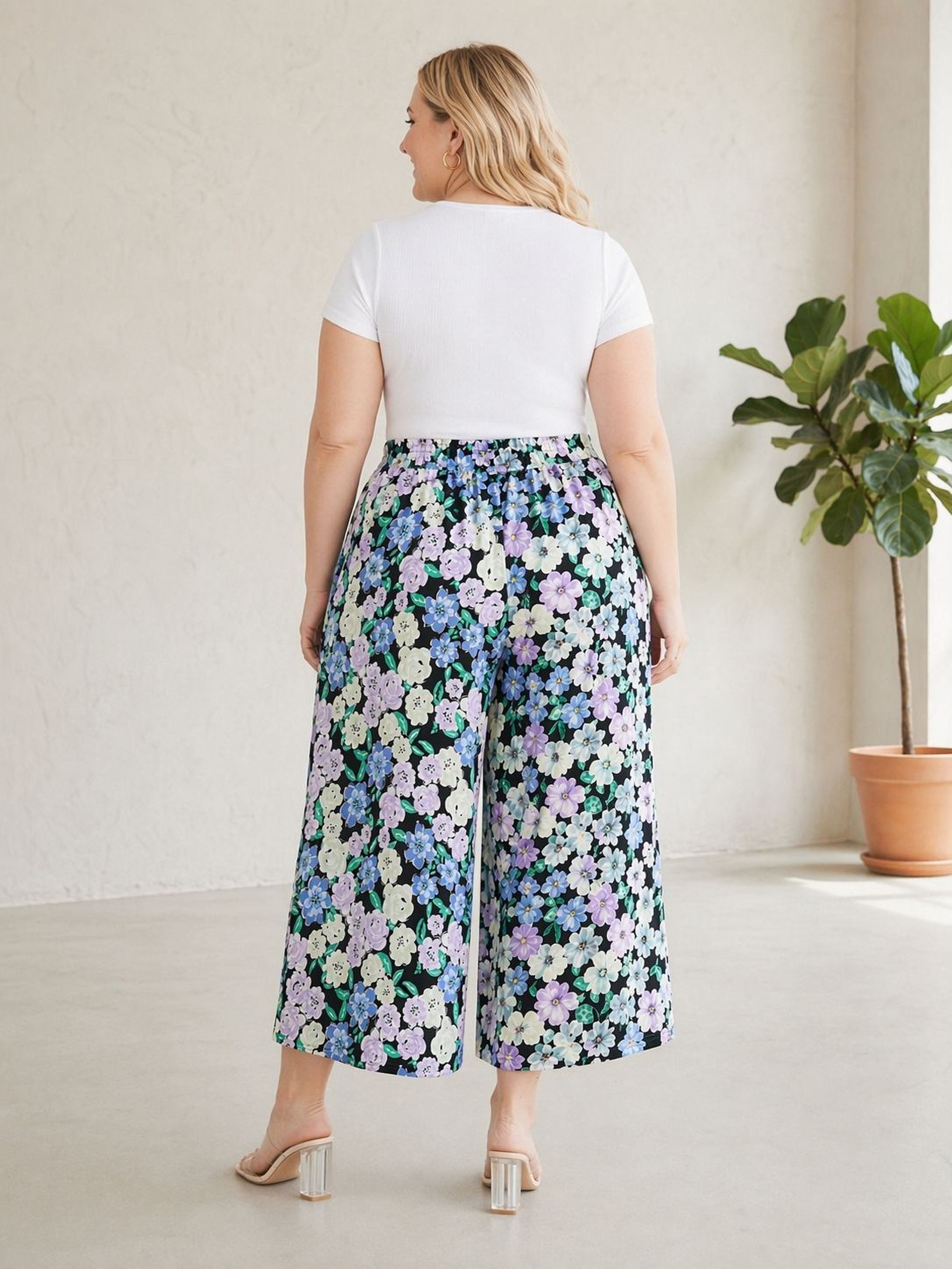 A woman wearing a white top and floral print wide-leg pants stands in a room with a potted plant.