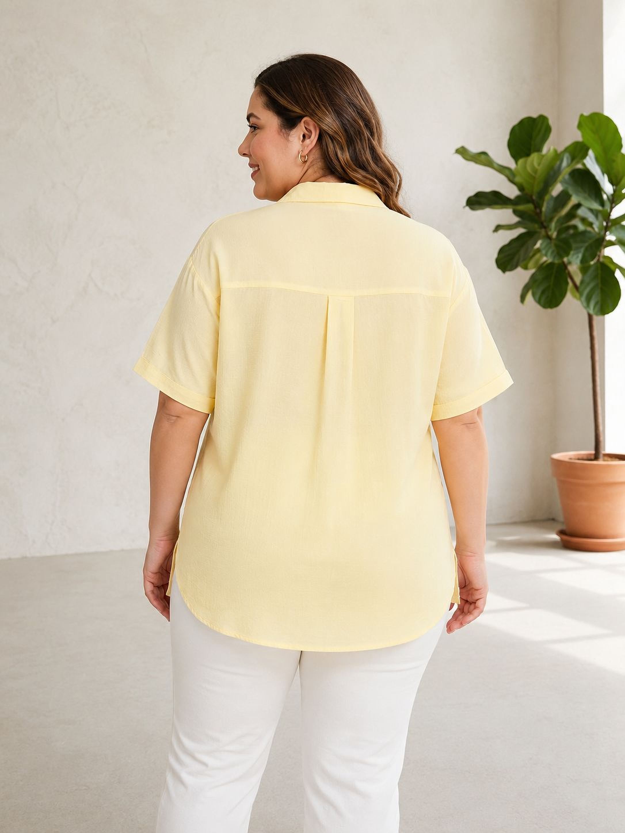 A woman wearing a yellow shirt and white pants stands in a room with a potted plant in the background.