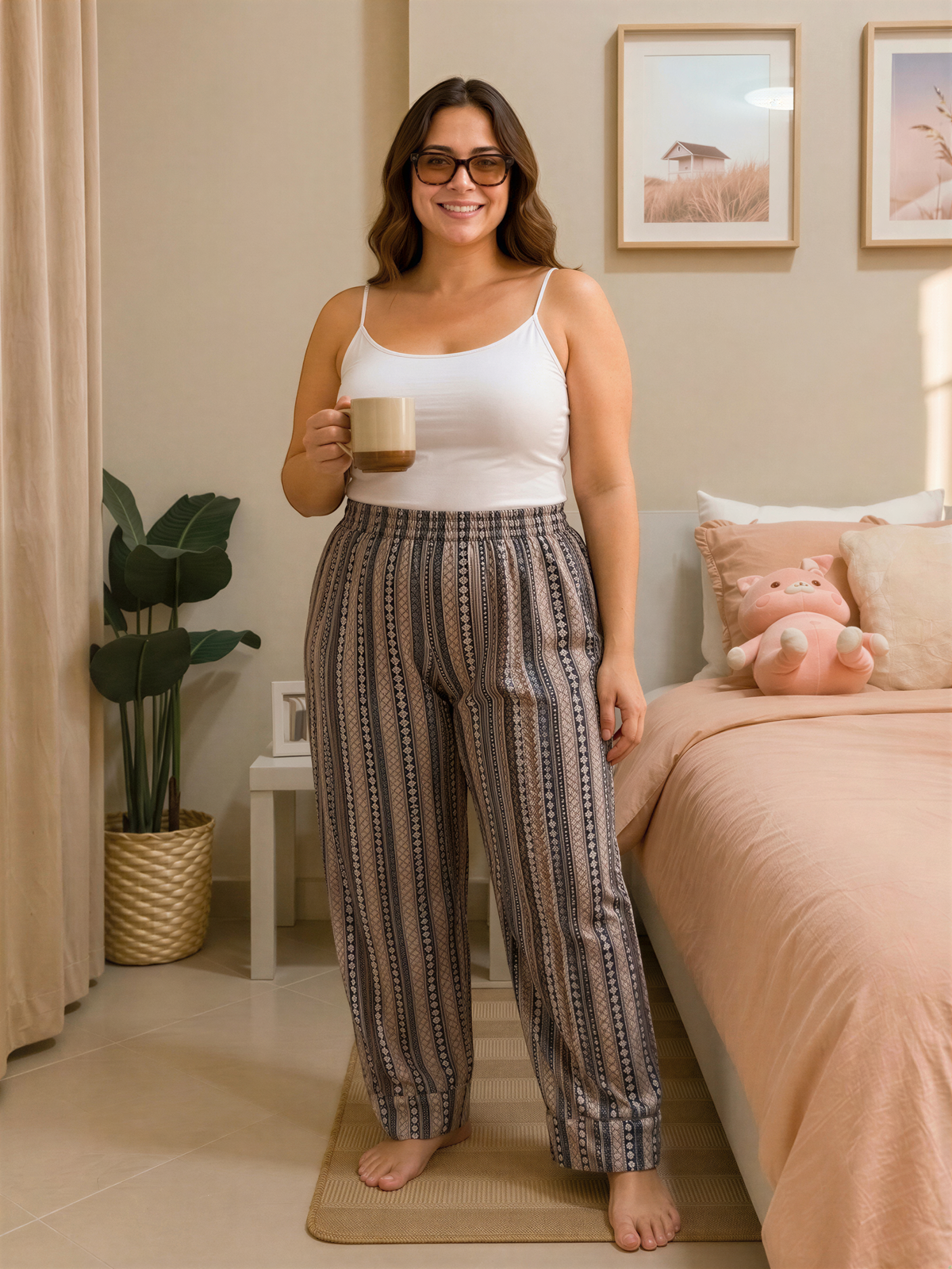 A woman in a white tank top and striped pants stands in a bedroom, holding a coffee mug and smiling.