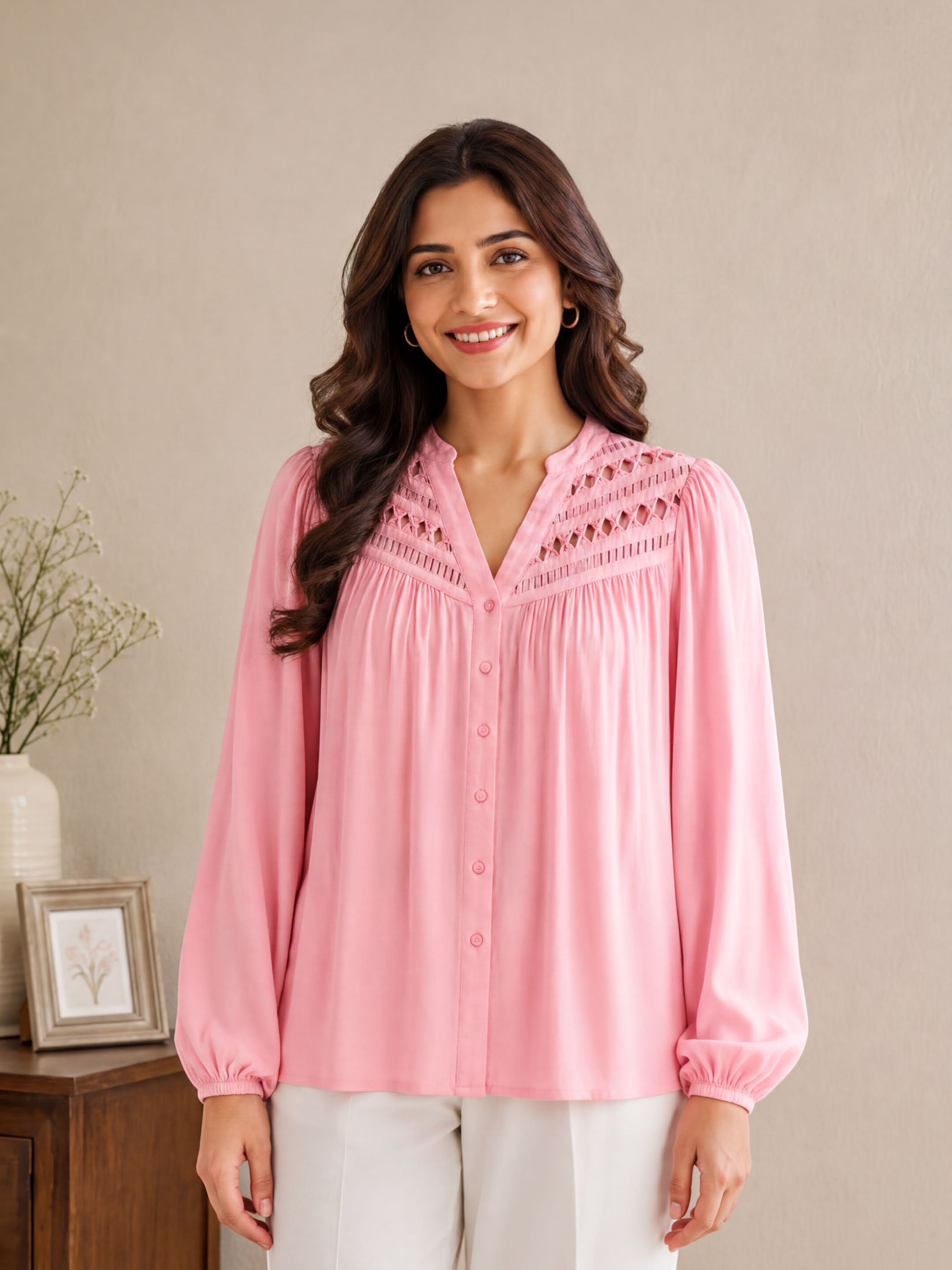 A woman with long brown hair wearing a pink blouse with a v neck and puffed sleeves stands in front of a wooden dresser with a picture frame on it.