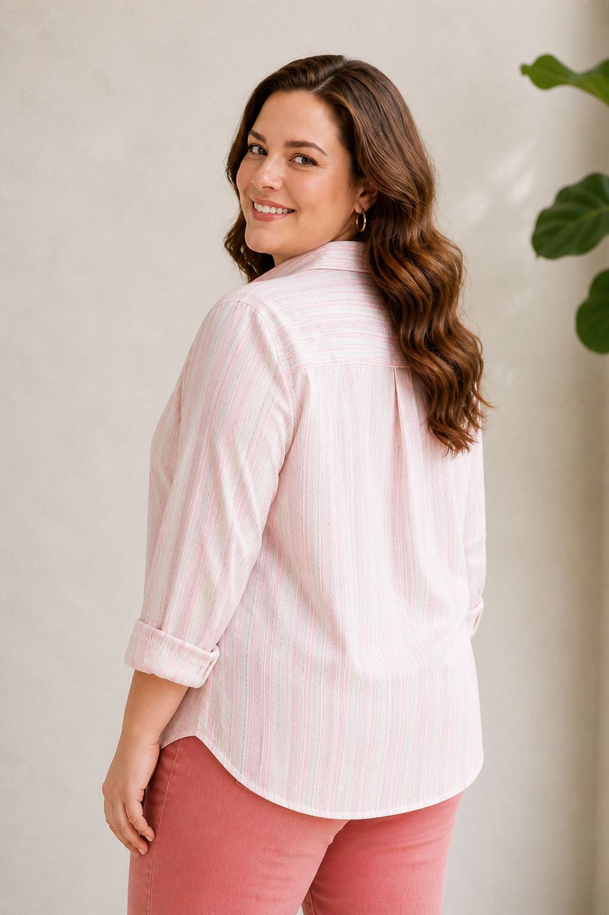 A woman with long brown hair is smiling and posing in a pink striped shirt and red pants against a plain background.