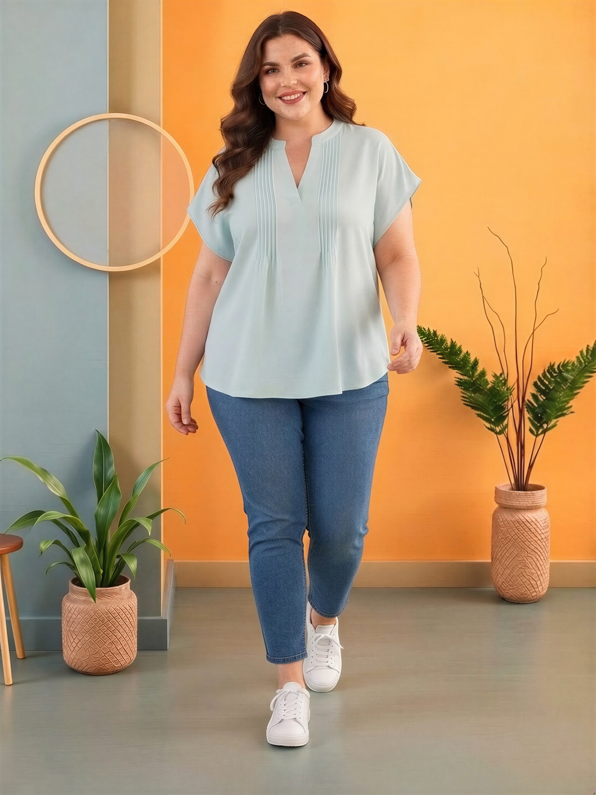 A woman wearing a light blue blouse and blue jeans is standing in a room with a wooden floor, a circular mirror, and two potted plants.