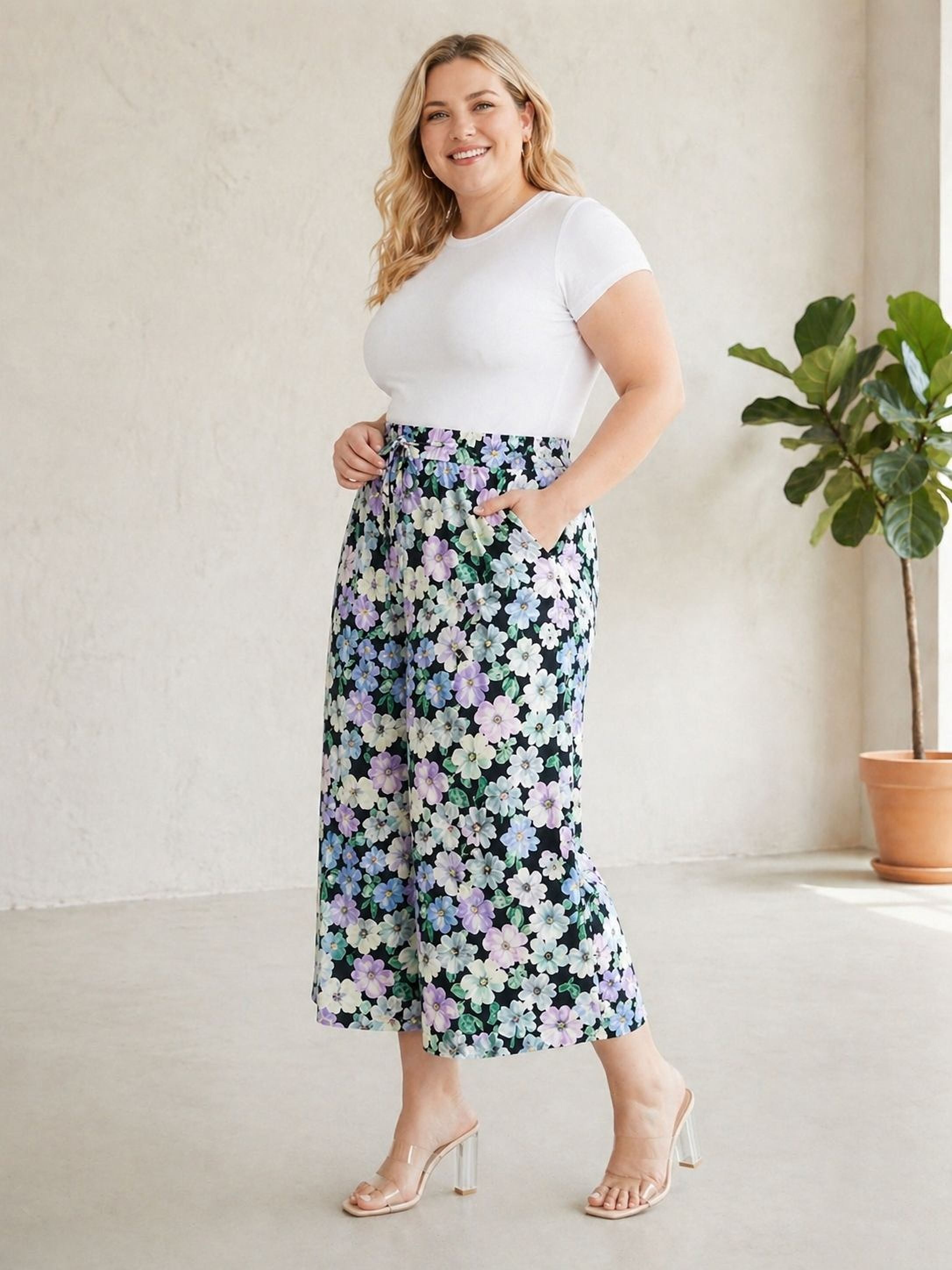 A woman wearing a white top and a floral print skirt is standing in a room with a potted plant in the background.