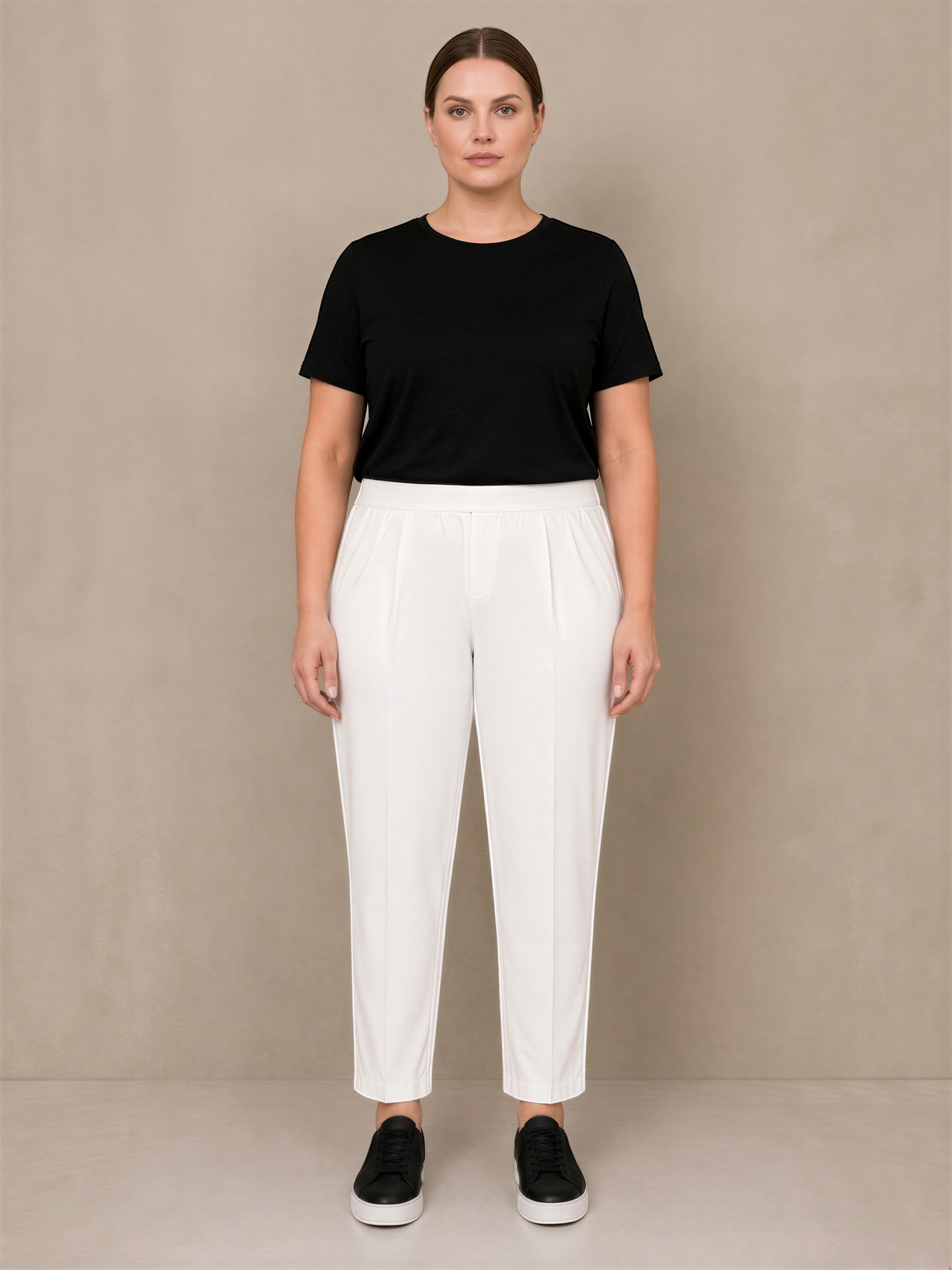 A woman wearing a black top and white pants stands against a beige background.