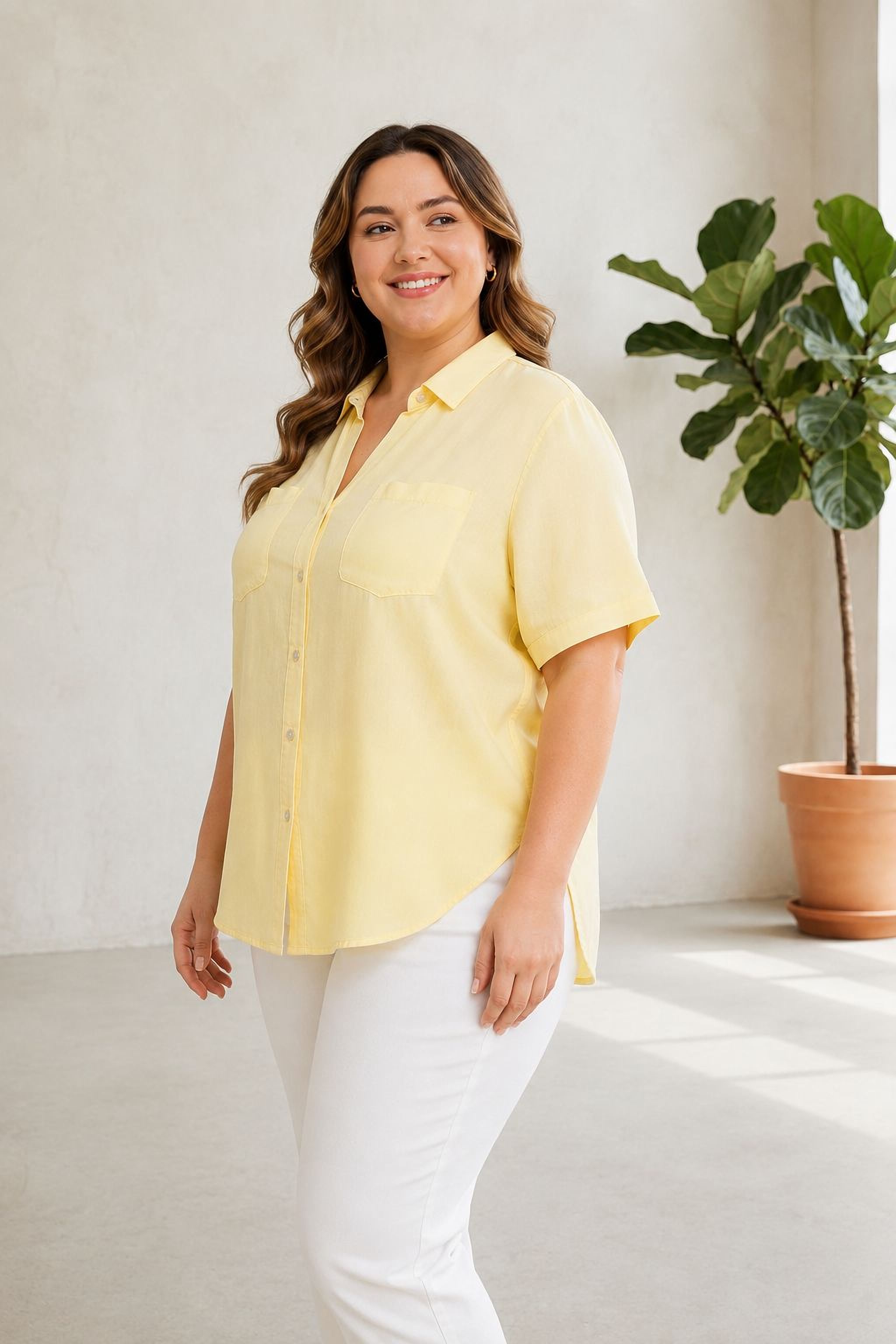A woman in a yellow shirt and white pants stands in a room with a potted plant in the background.